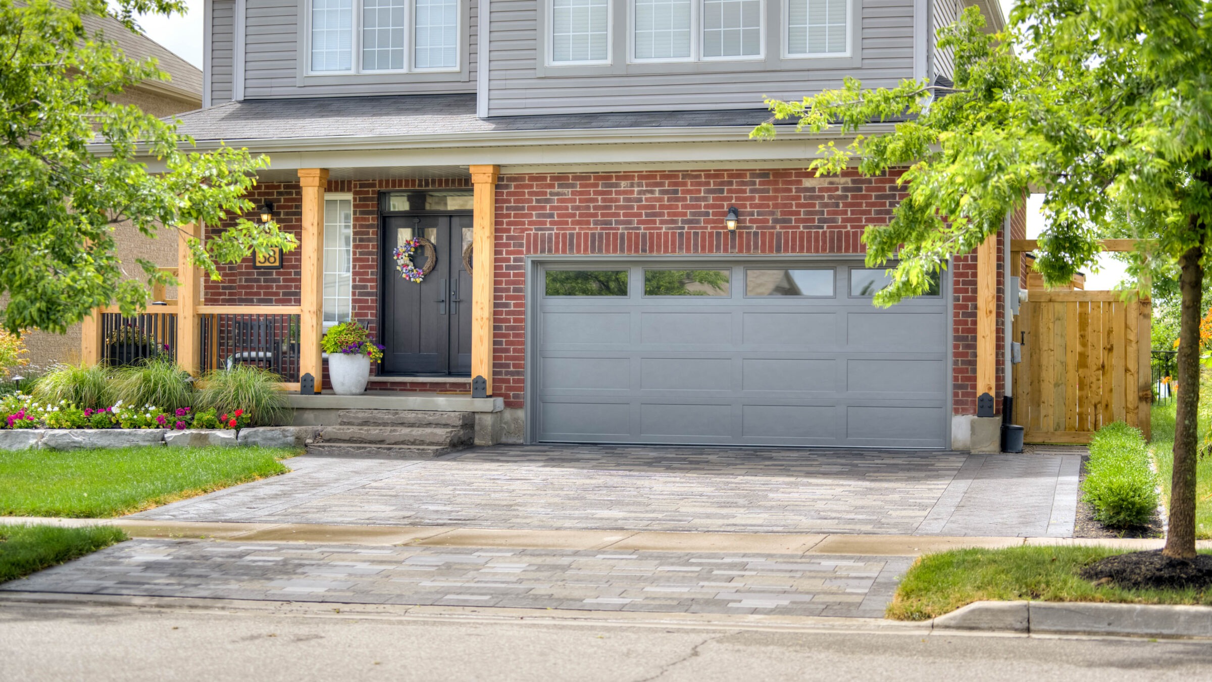 A modern suburban house with a brick facade, gray garage, and decorative wreath. Manicured lawn and trees enhance the pleasant, inviting atmosphere.