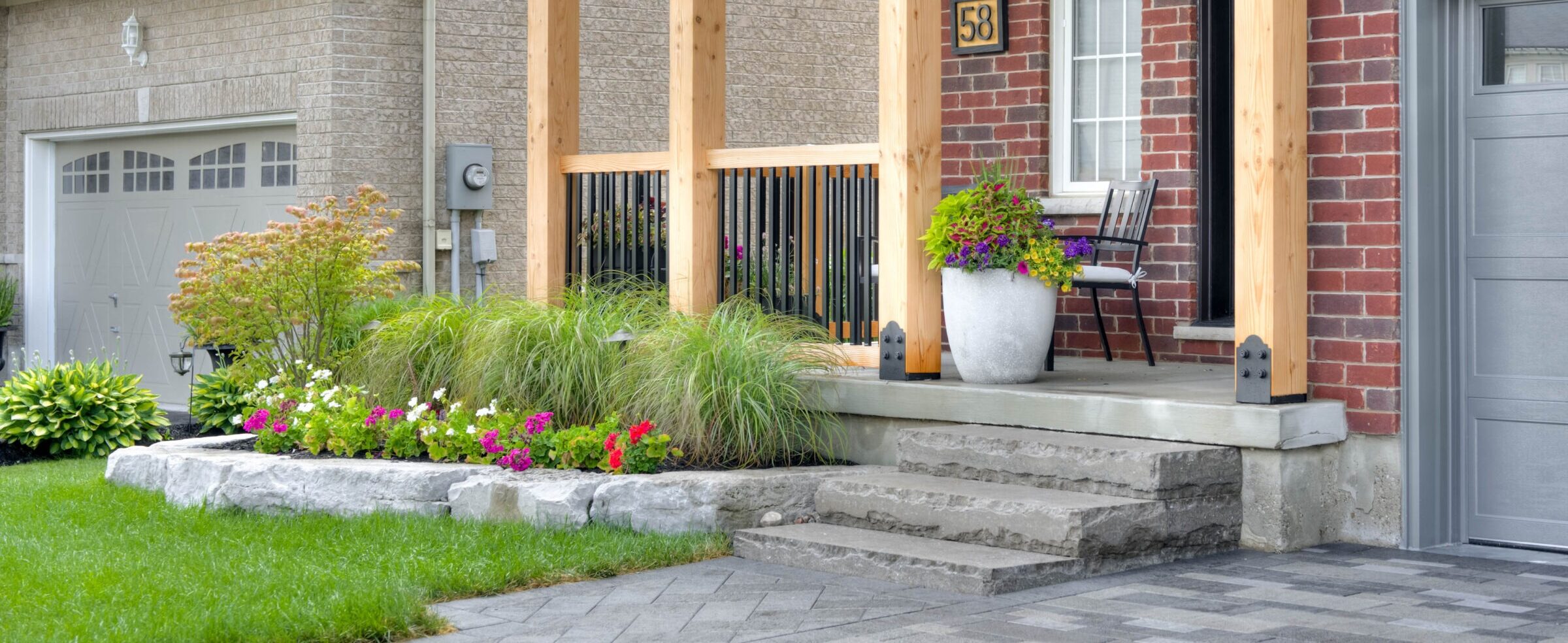 Brick house exterior with wooden porch, colorful potted flowers, and stone steps. Well-maintained garden surrounds entrance, creating a welcoming home atmosphere.