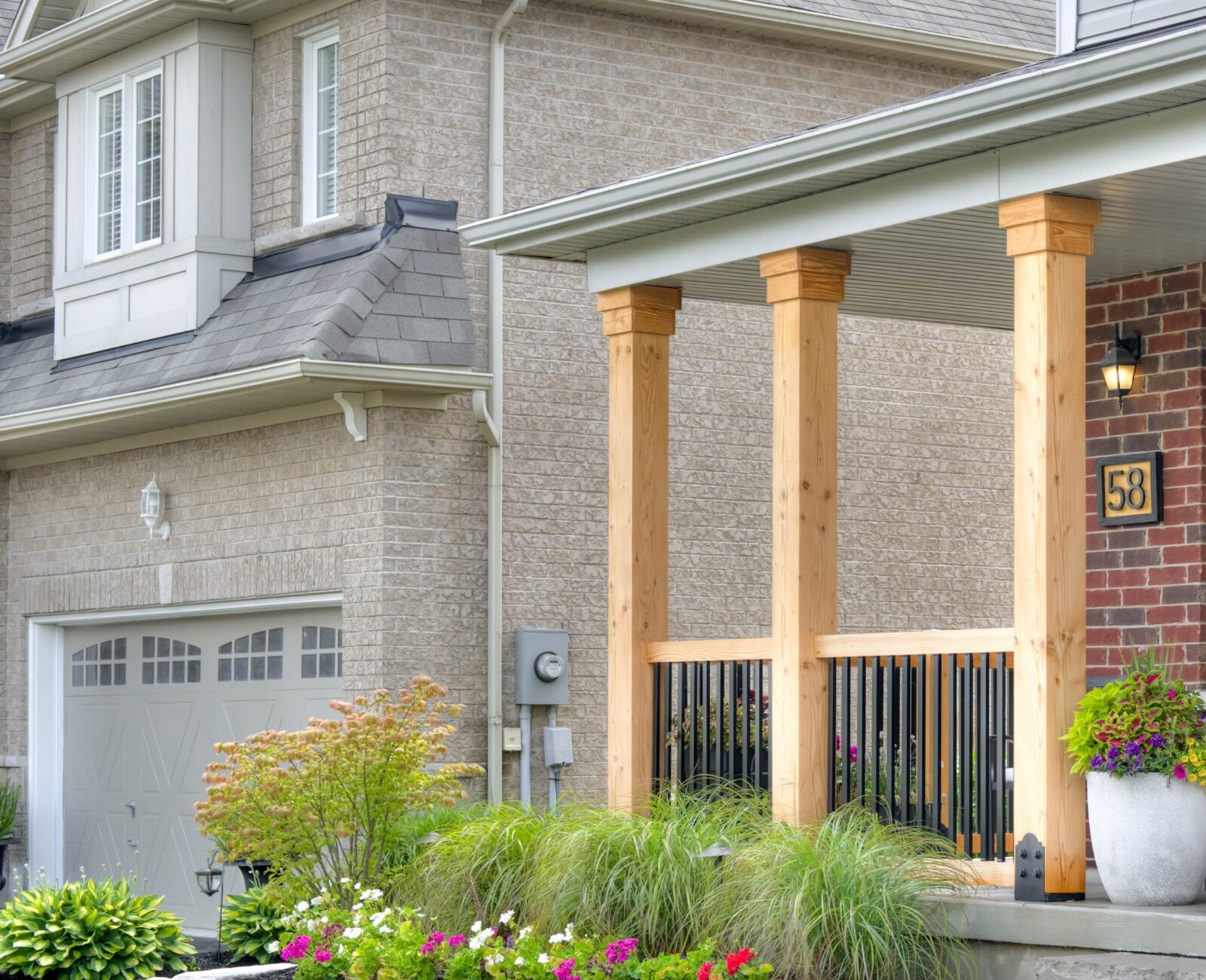 Brick house with wooden porch, potted flowers, and chair. Well-kept garden surrounds stone steps. Modern suburban home with attached garage and house number.