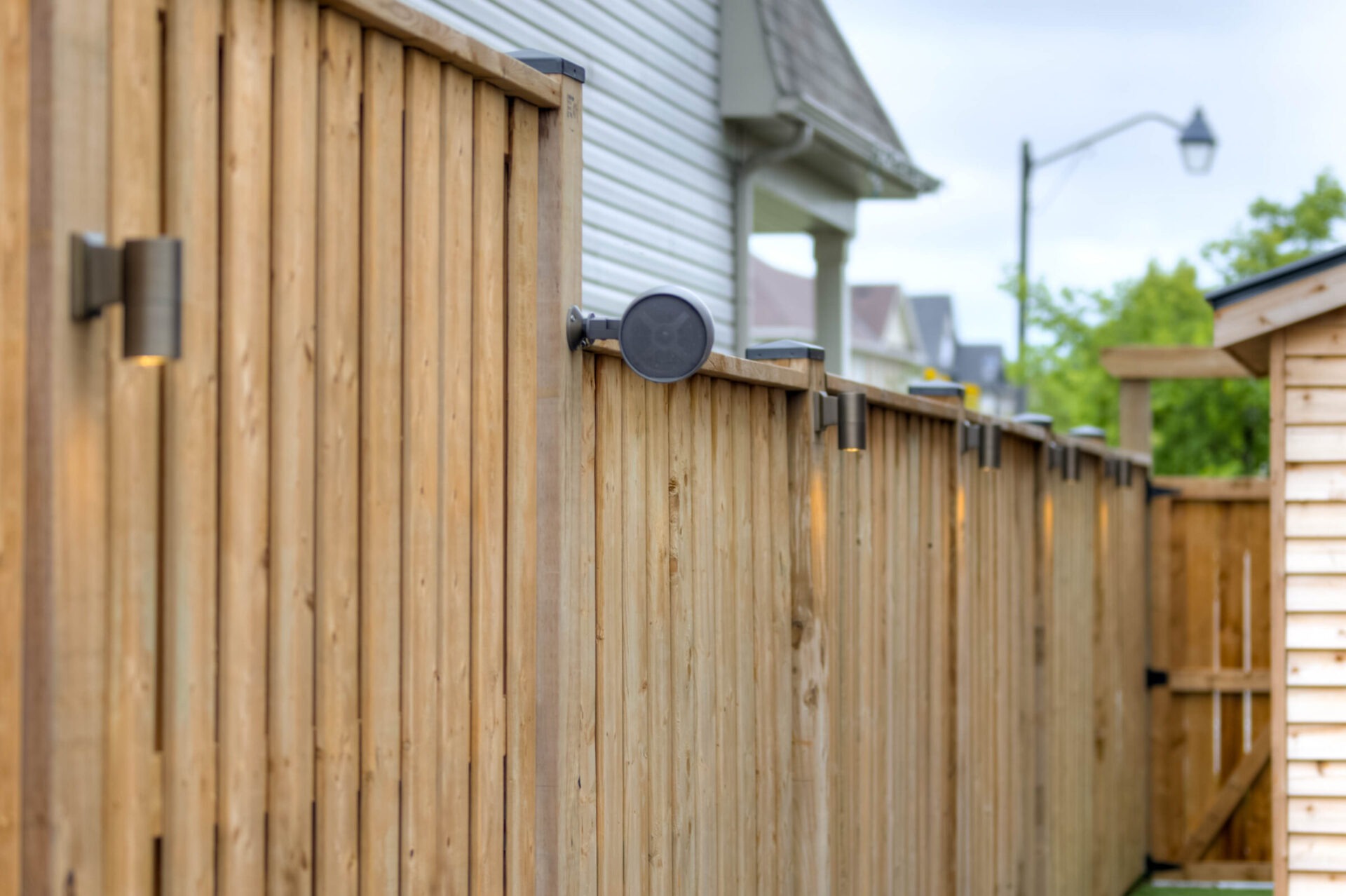 Wooden fence with mounted lights and outdoor speaker, beside a white house. The scene captures a residential neighborhood setting with trees and sky visible.