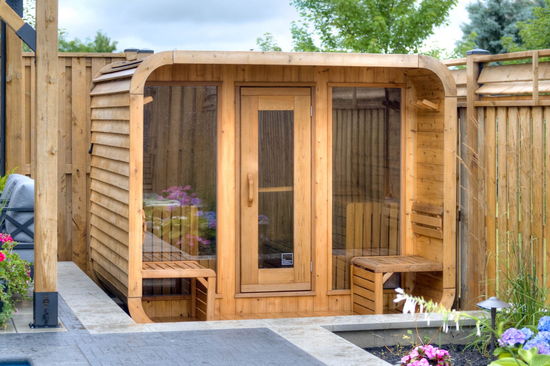 Wooden outdoor sauna with a glass door, surrounded by a wooden fence. Colorful flowers and greenery enhance the peaceful backyard setting.