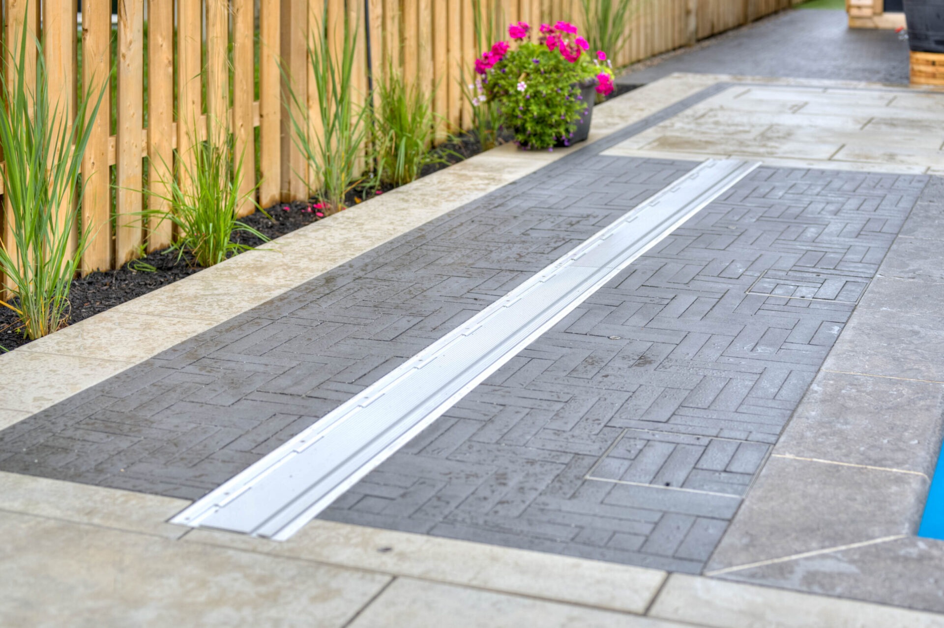 A landscaped patio features a wood fence, potted flowers, gray herringbone tiles, and a drainage channel amidst greenery, creating a serene outdoor space.