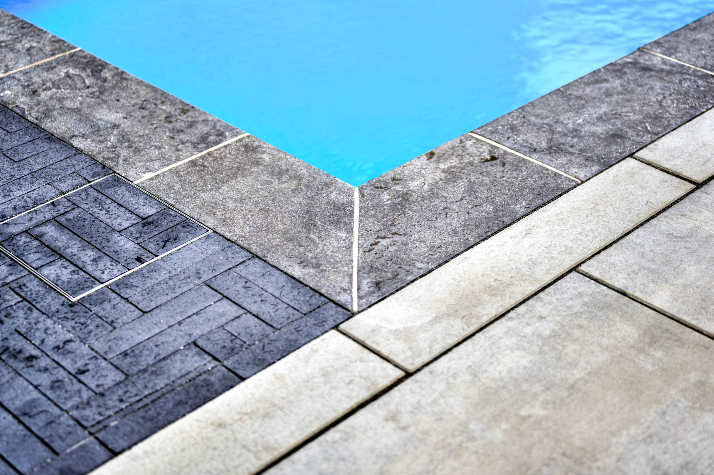 Pool edge with various textured tiles in gray and black, meeting clear blue water. Clean and modern design, no people visible.