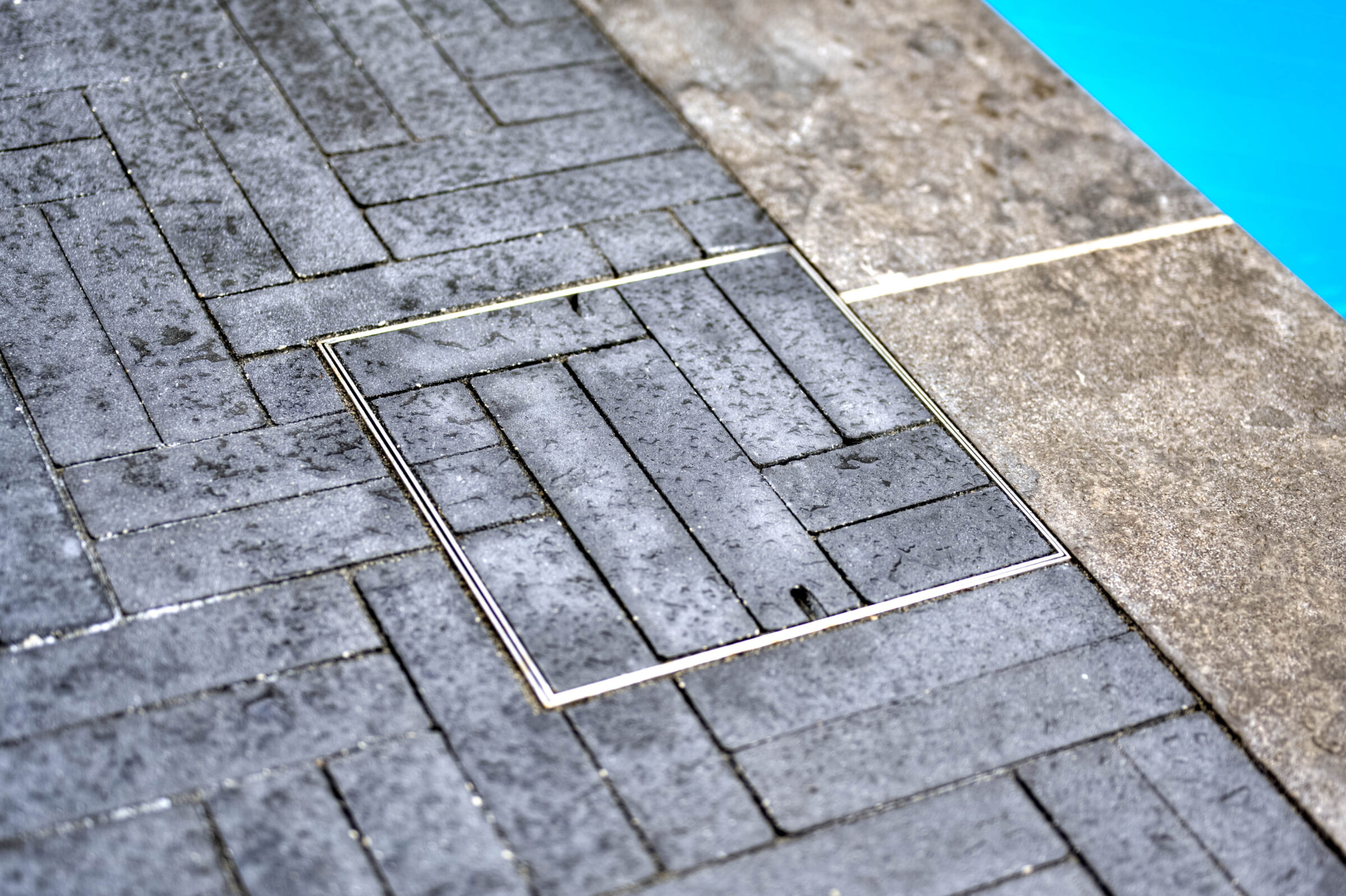 Water cascades onto a herringbone-patterned stone surface beside a pool, creating a serene and minimalist atmosphere. No landmarks visible.