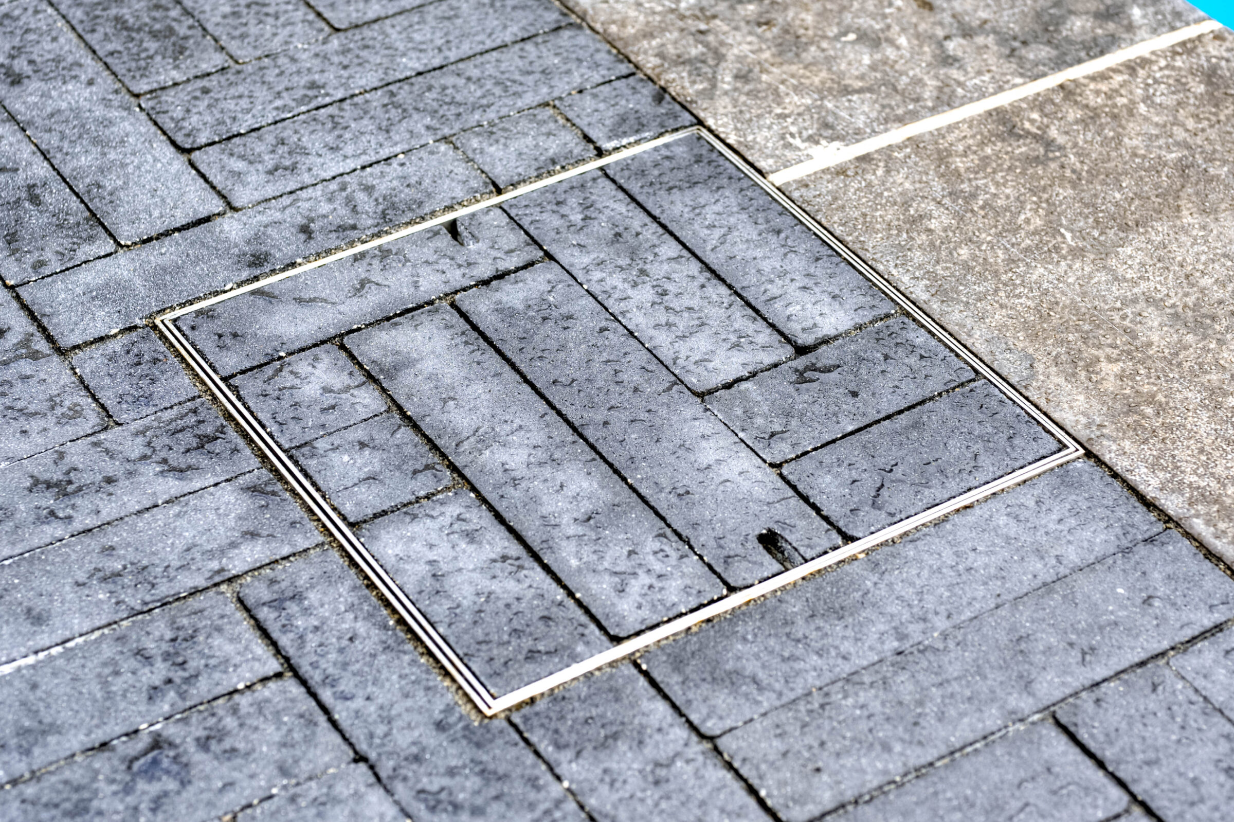 An image of a wet, grey brick pavement with a rectangular access panel blending seamlessly into the design, surrounded by smoother stone tiles.