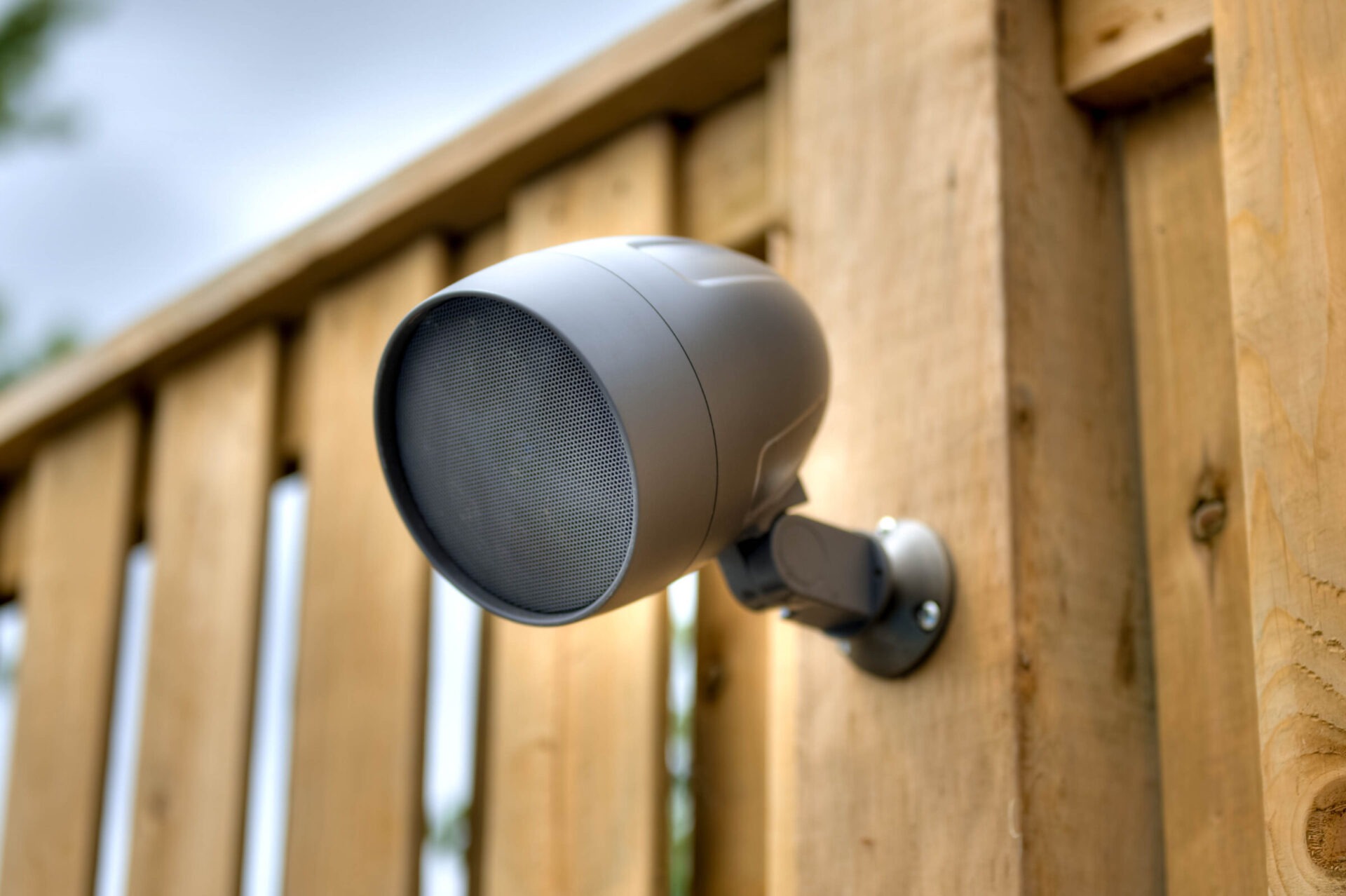 A gray outdoor speaker mounted on a wooden fence, set against a cloudy sky backdrop. The image emphasizes sound equipment integration into environments.