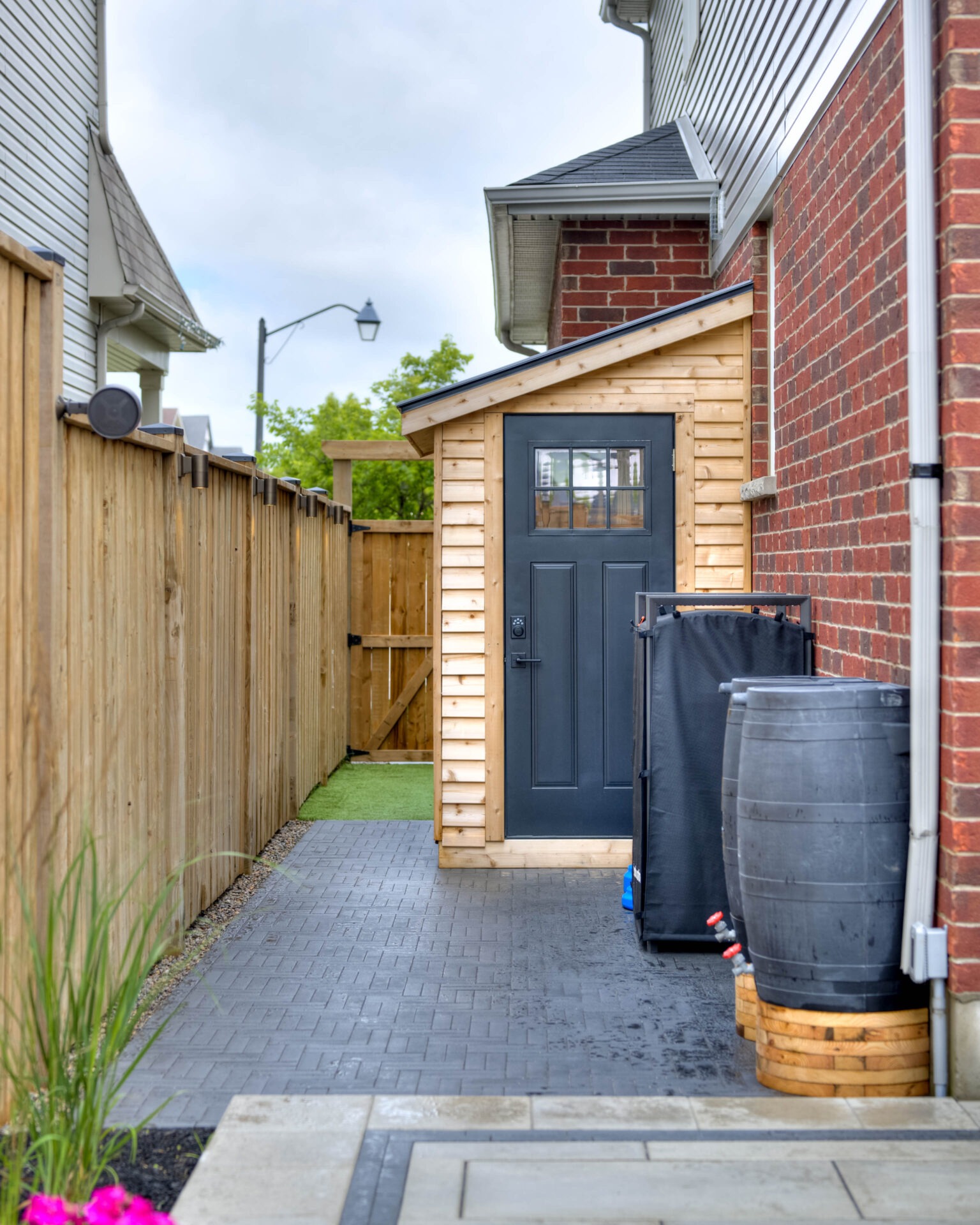 A tidy backyard features a small wooden shed with a black door, rain barrels, and a wooden fence under a cloudy sky.