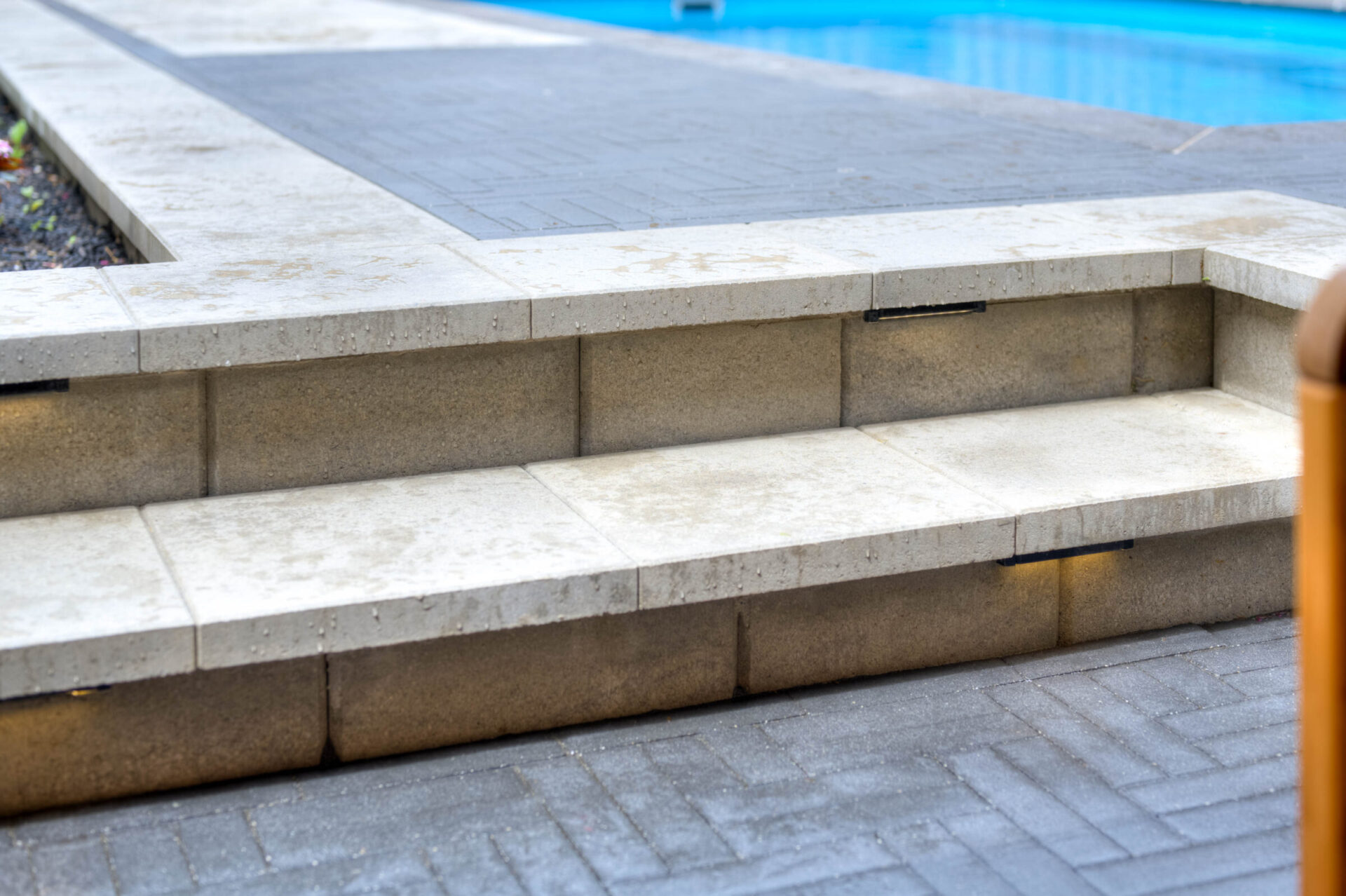Concrete steps lead to paved walkway beside a pool. Subtle LED lights illuminate the stair edges, providing modern outdoor ambiance.