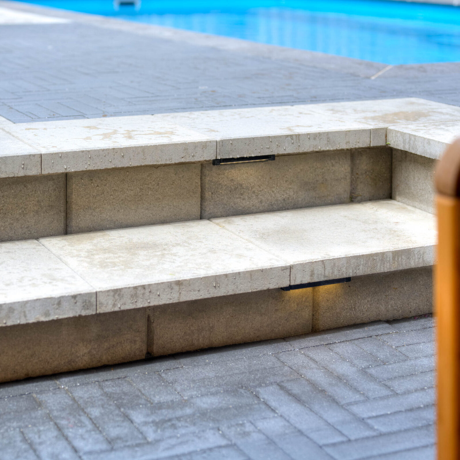 Concrete steps lead to paved walkway beside a pool. Subtle LED lights illuminate the stair edges, providing modern outdoor ambiance.