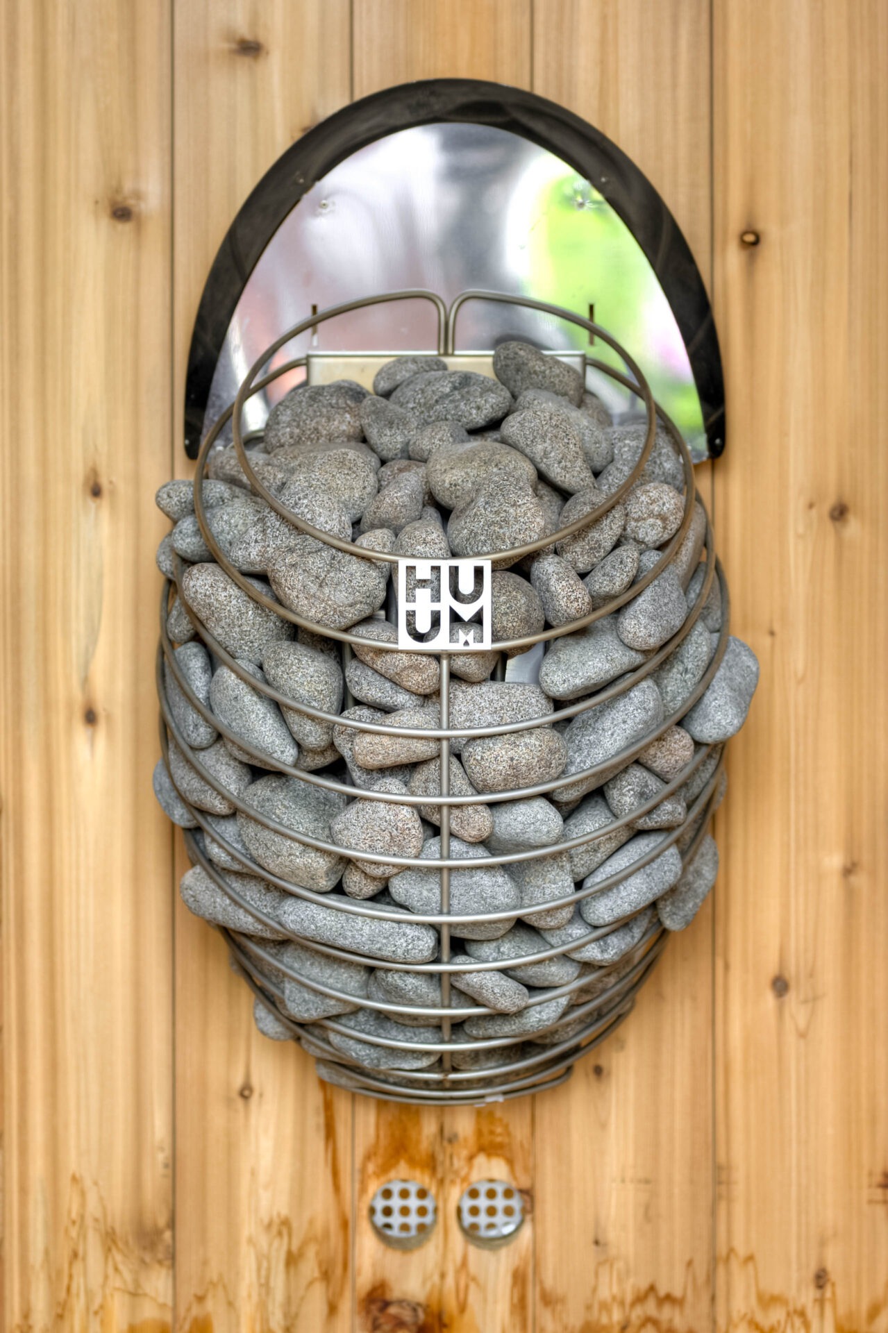 A wall-mounted sauna heater with stones inside a metal basket on a wooden surface, featuring the brand name HUUM prominently displayed.