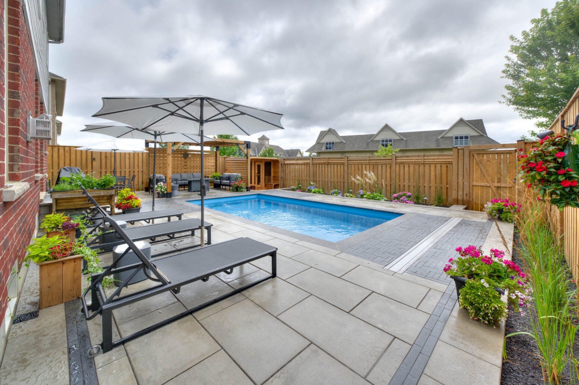 A modern backyard with a rectangular pool, sun loungers, umbrellas, and vibrant flowers, all enclosed by a wooden fence and lush greenery.