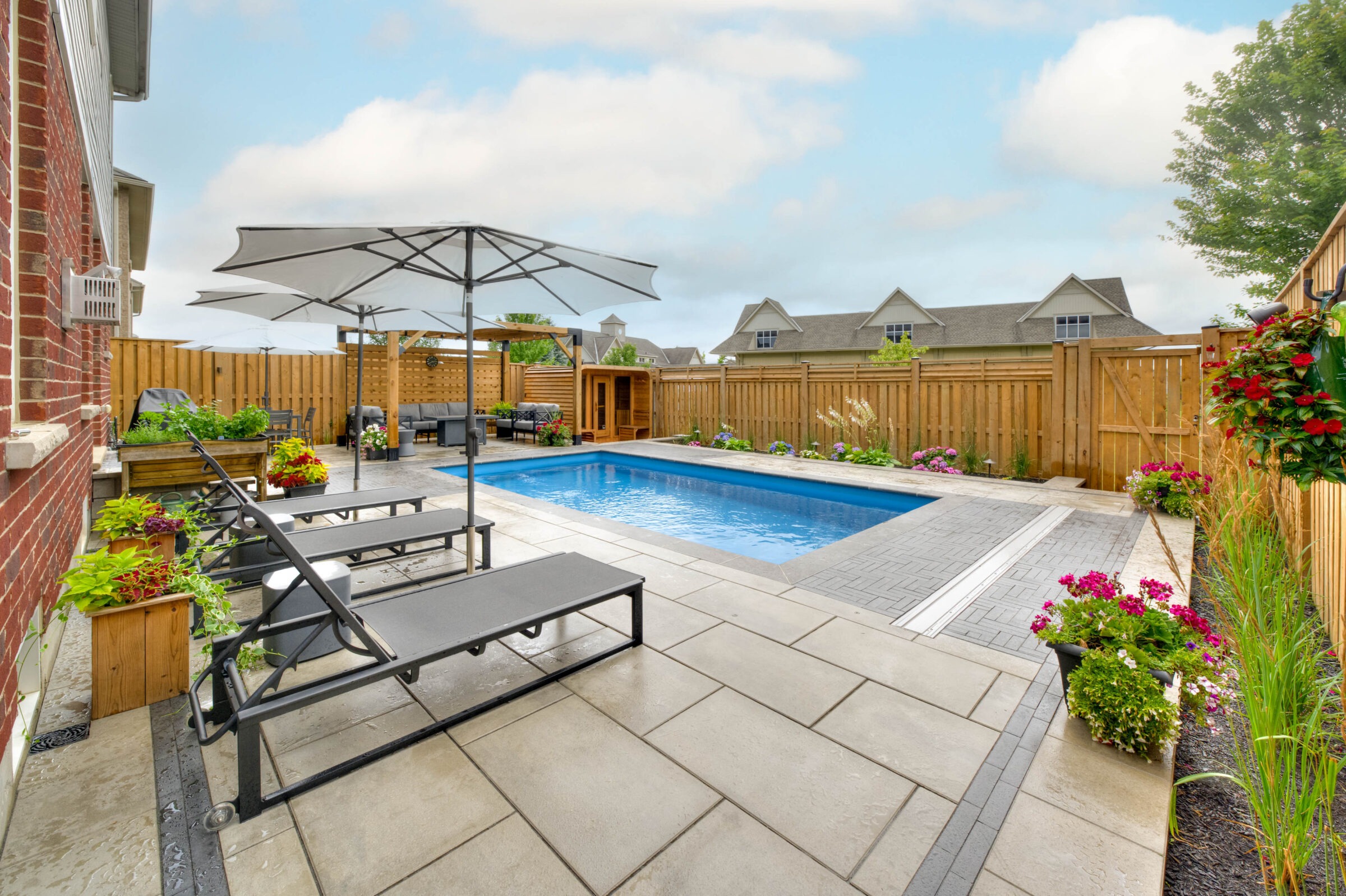 A private backyard features a rectangular pool, surrounded by lounge chairs, potted plants, and a wooden fence under a clear blue sky.