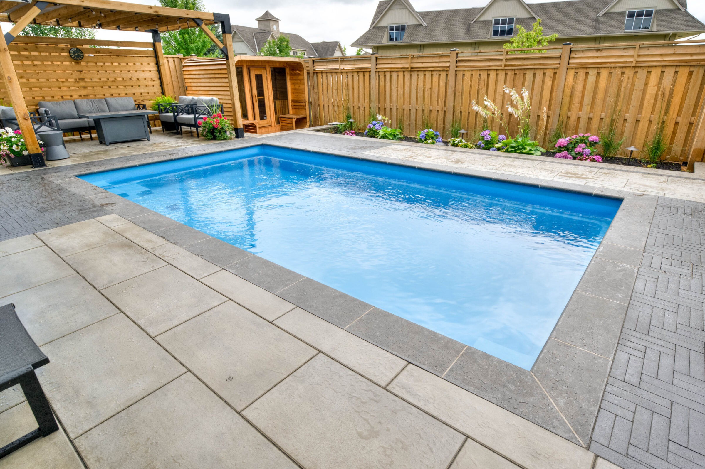 A small backyard features a rectangular pool, wooden fence, pergola, gray seating, and flowering plants, creating a relaxing outdoor retreat.