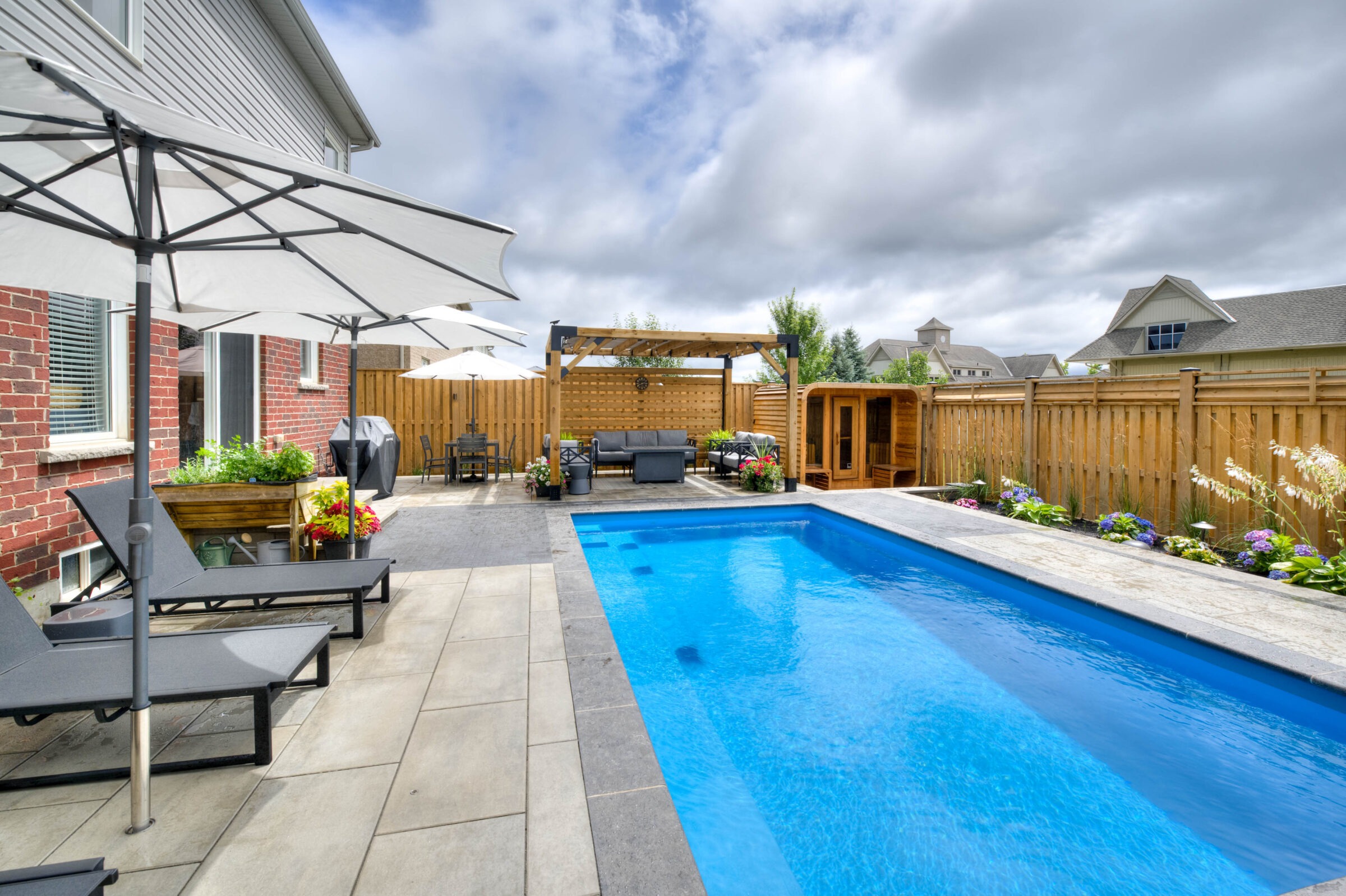 A serene backyard featuring a swimming pool, patio furniture, wooden pergola, and vibrant garden, surrounded by a clean, wooden fence under a cloudy sky.