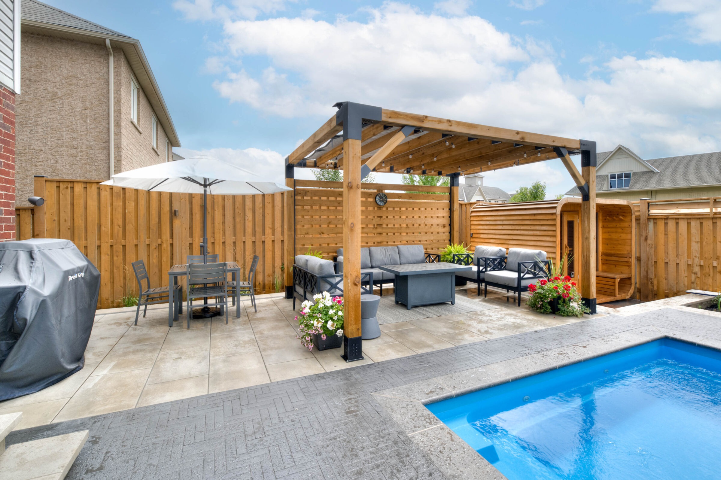 A backyard with a pool, wooden pergola, outdoor furniture, a grill, and a dining area under an umbrella, surrounded by a fence.