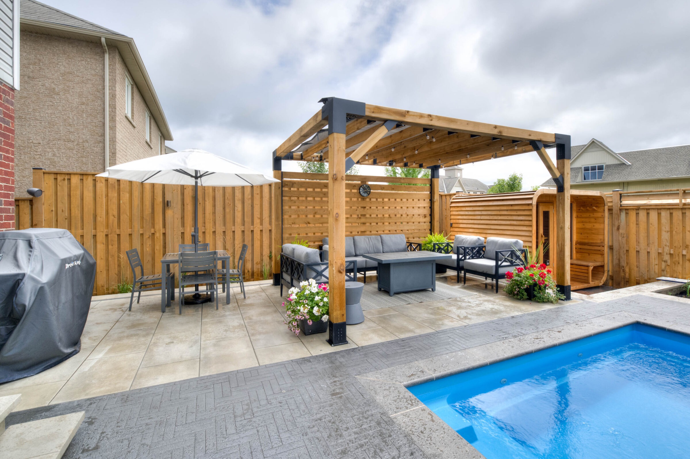 A backyard features a pergola, cozy seating, table with umbrella, sauna, grill, blue pool, and vibrant flowers, surrounded by wooden fencing.