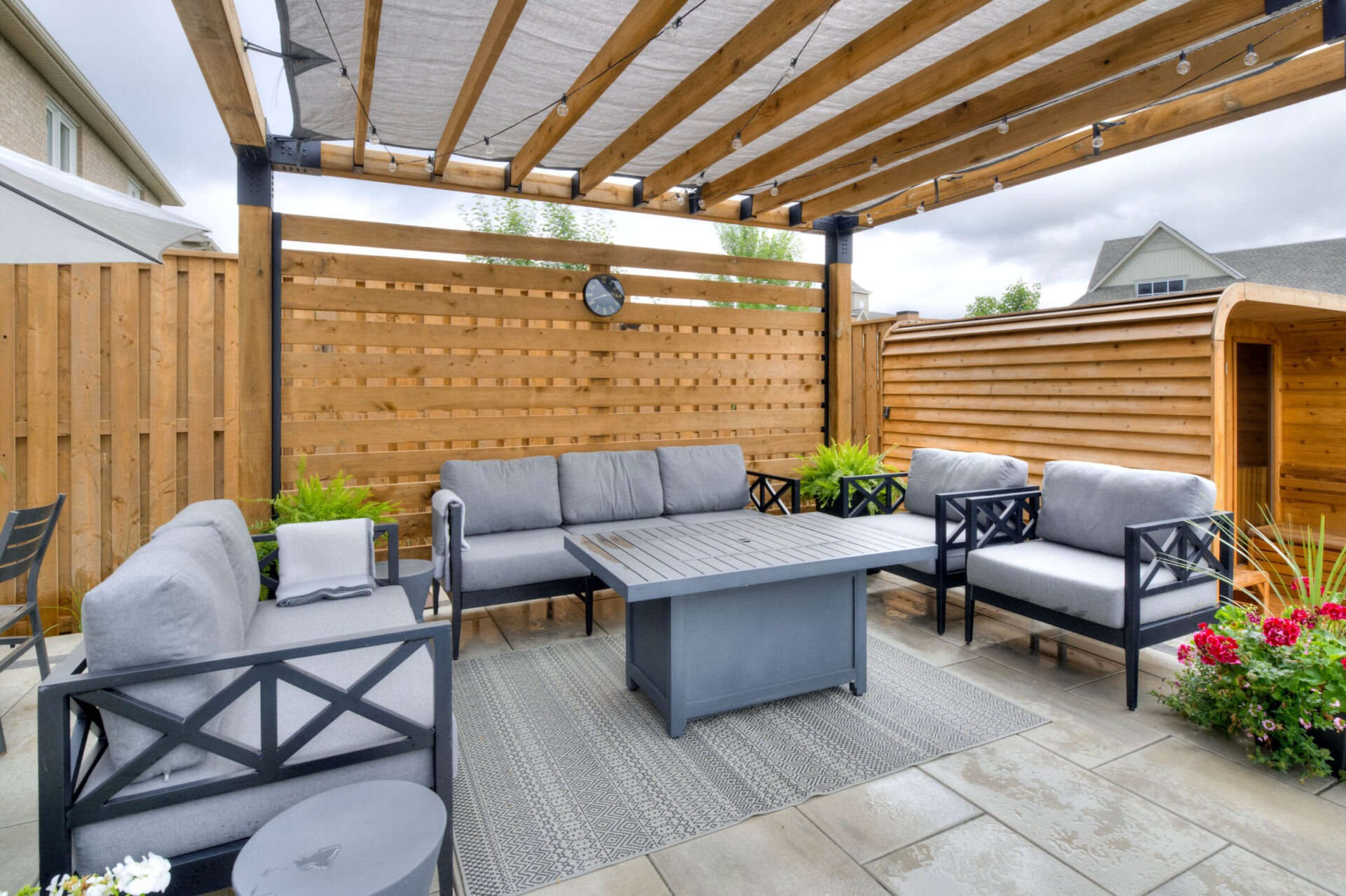 A modern outdoor patio with wooden pergola, gray seating, a central table, and surrounding plants, creating a cozy relaxation space.