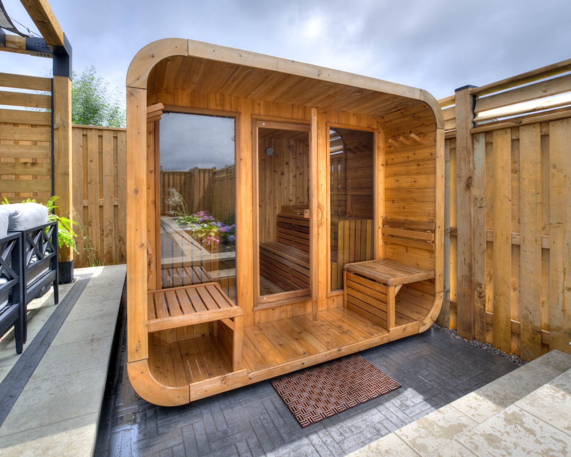 Modern wooden sauna with glass door, surrounded by a wooden fence and patio furniture. The space is adorned with plants and flowers.