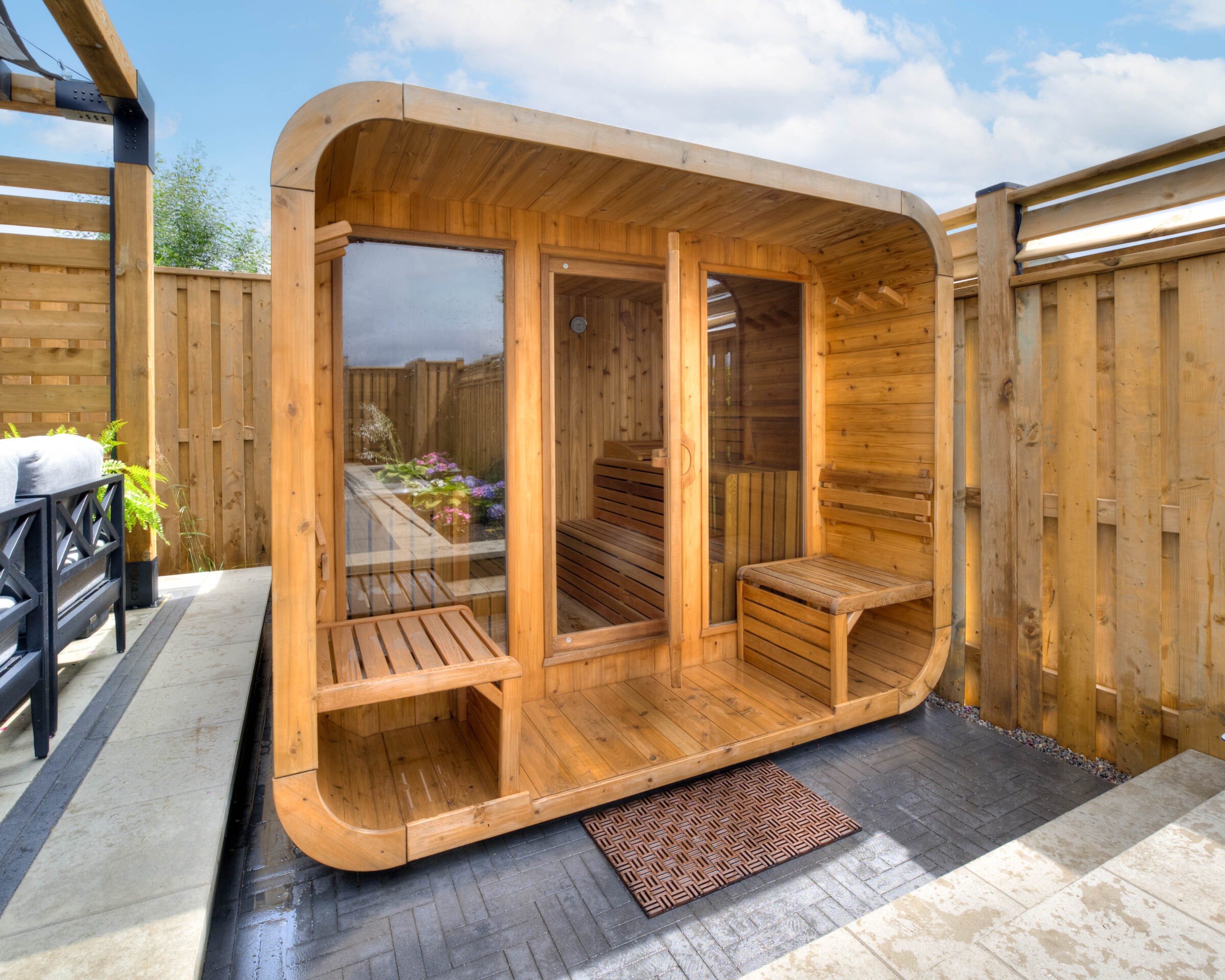 A modern, small wooden sauna is situated on a patio, surrounded by a wooden fence, with patio furniture nearby and clear skies above.