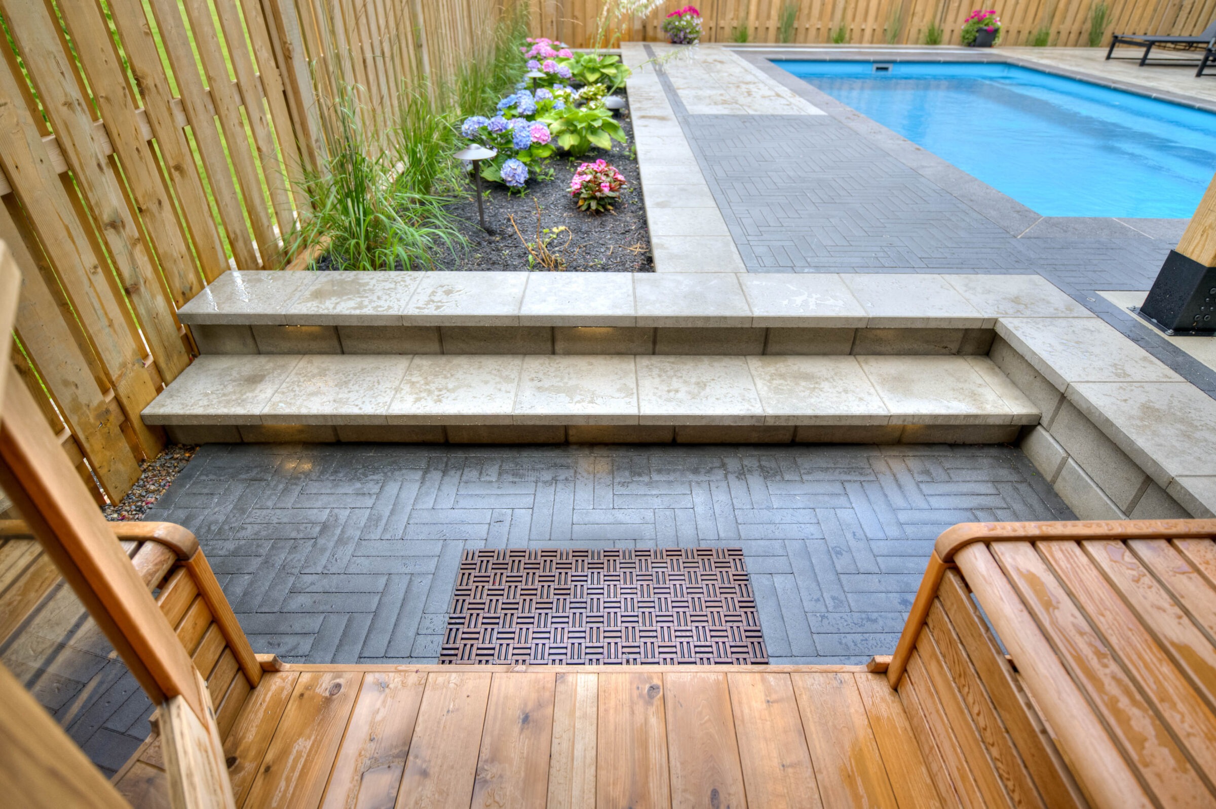 A backyard features a modern pool with paver steps, a wooden deck, colorful flowers, and a wooden fence surrounding the area.