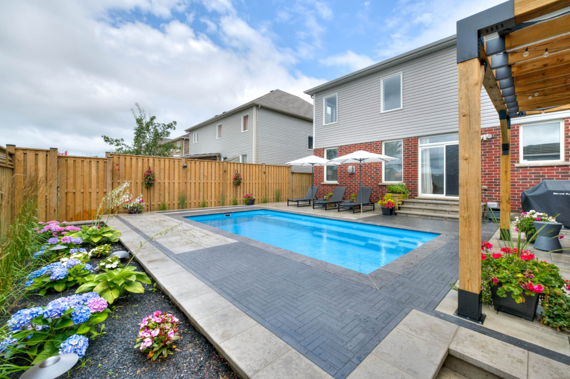 A backyard features a blue swimming pool, surrounded by colorful flowers, patio chairs, umbrellas, a wooden fence, and adjacent brick house.