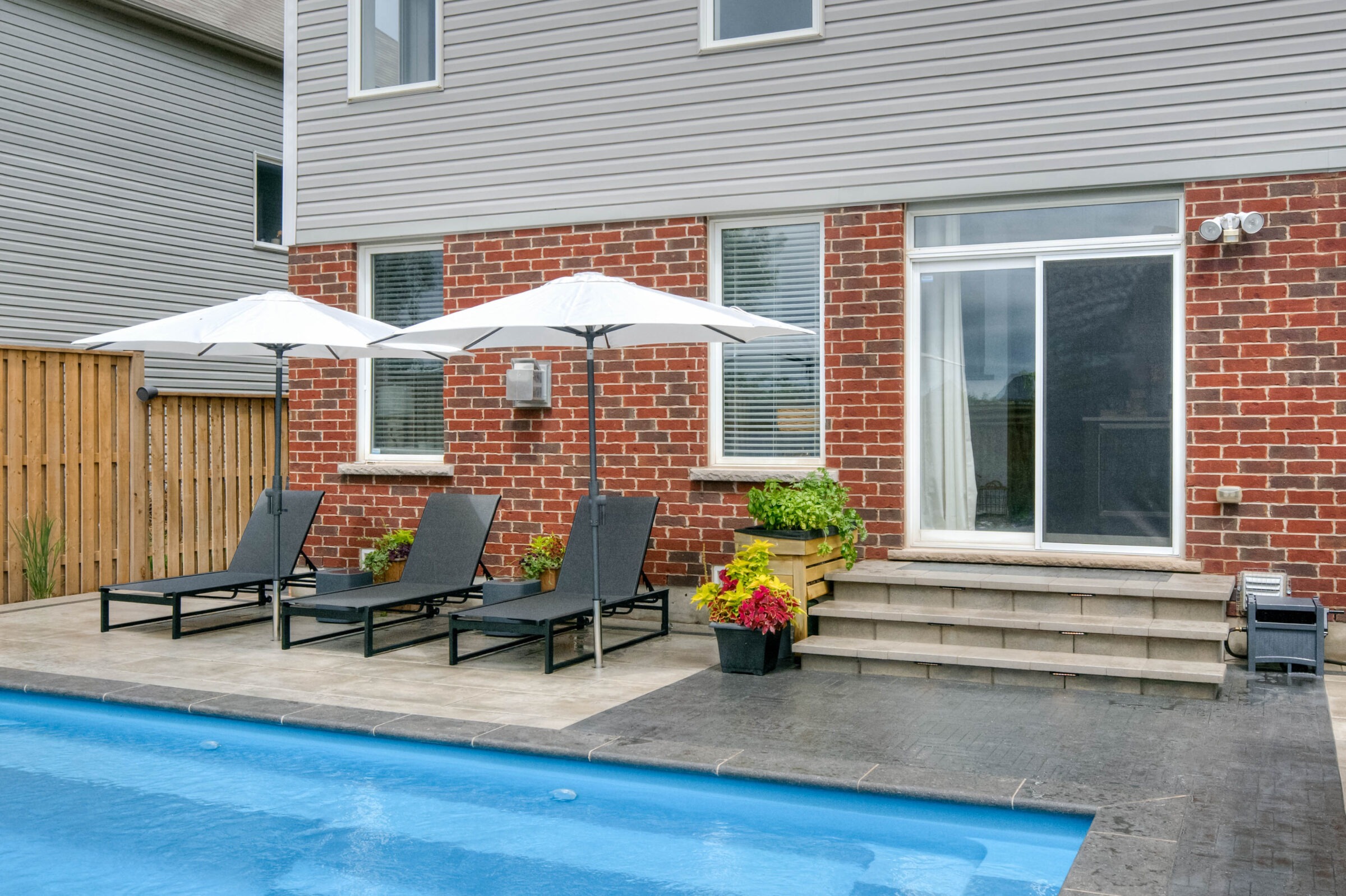 Brick house with sliding glass door, poolside loungers, and umbrellas. Wooden fence, potted plants, and steps leading to a patio area.