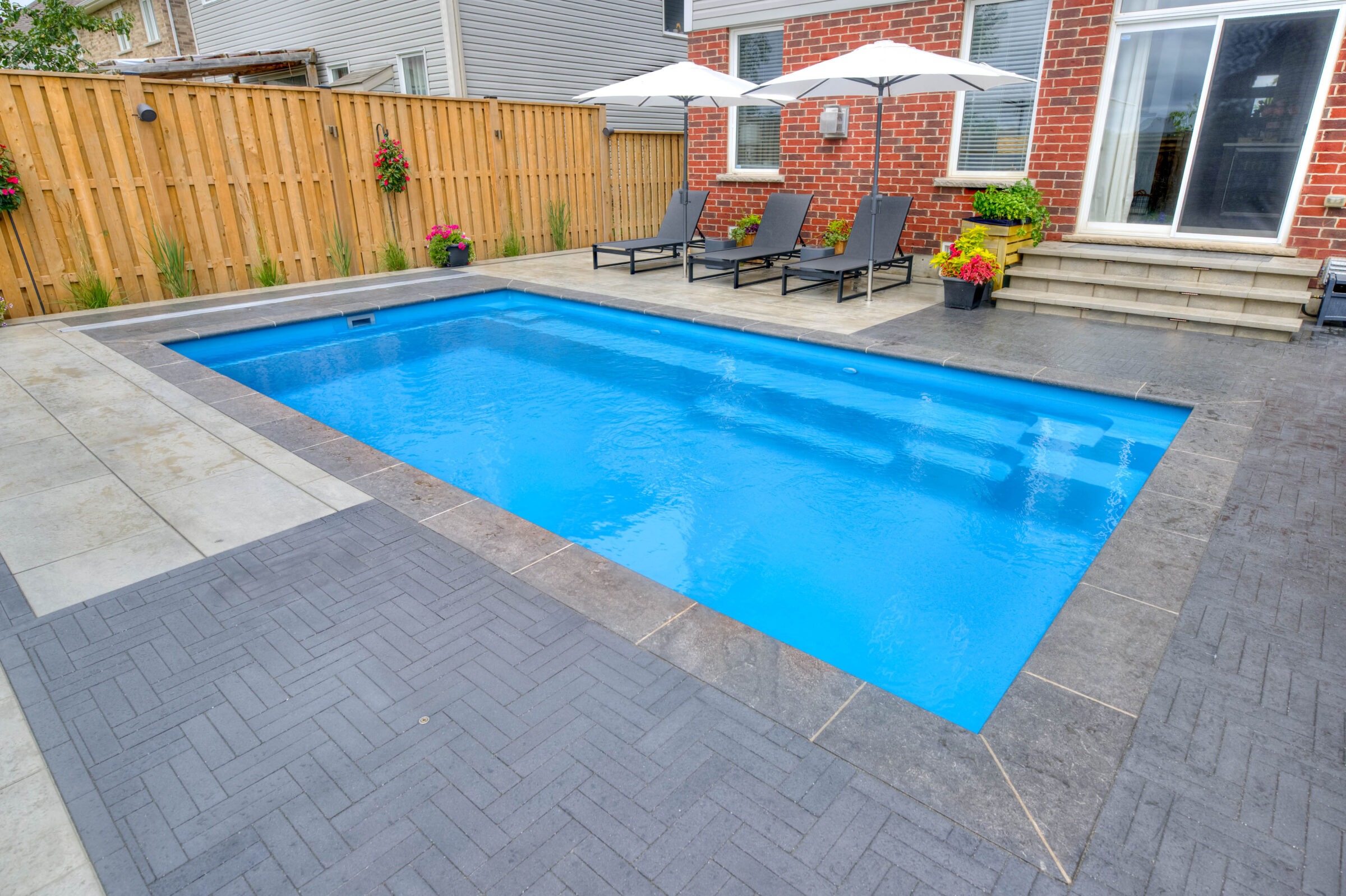 A backyard features a rectangular swimming pool, sun loungers under umbrellas, and potted plants along a wooden fence and brick house.