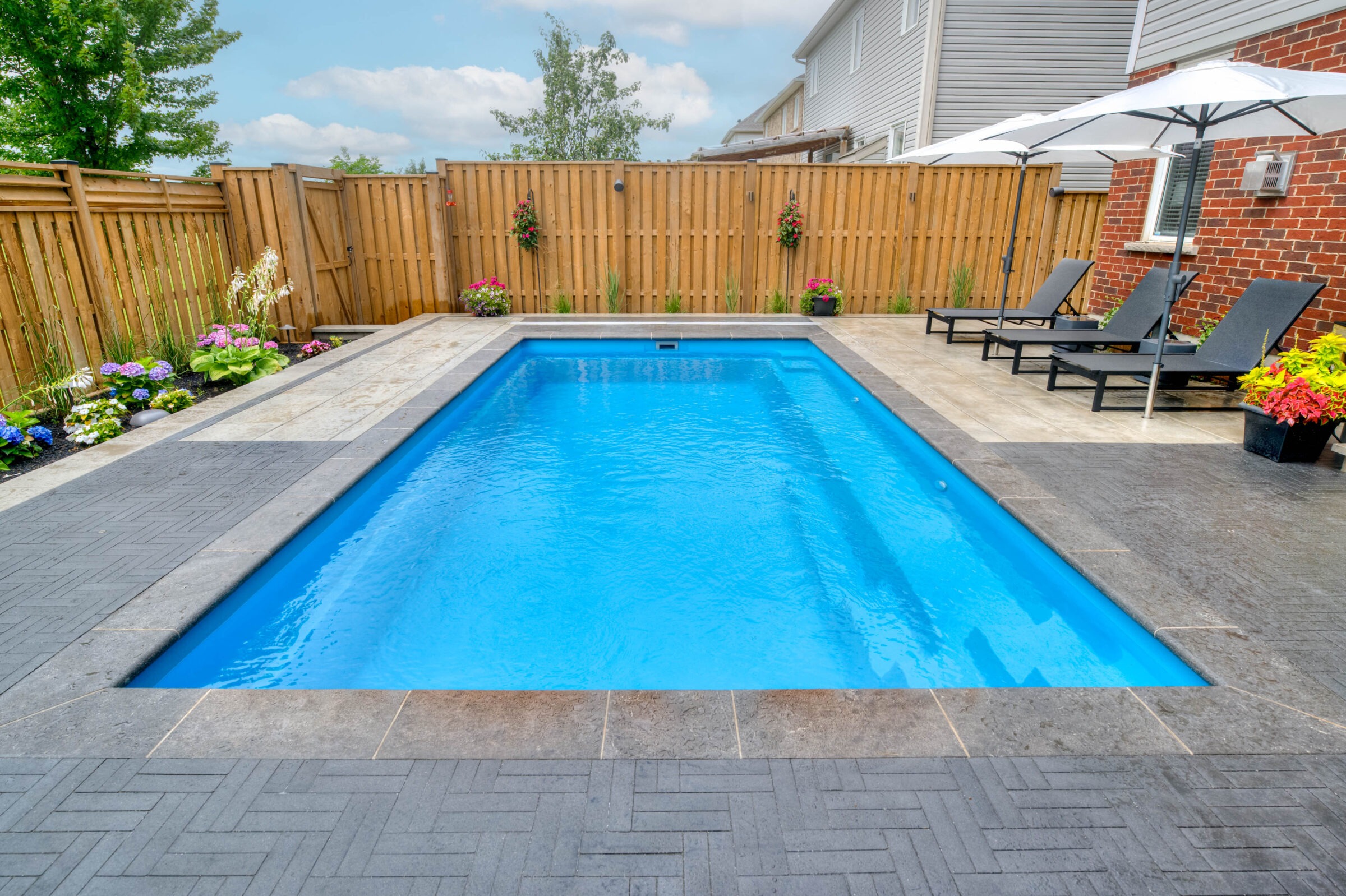 A backyard swimming pool with lounge chairs, umbrella, and vibrant flowers, surrounded by a wooden fence and adjacent to a brick house.