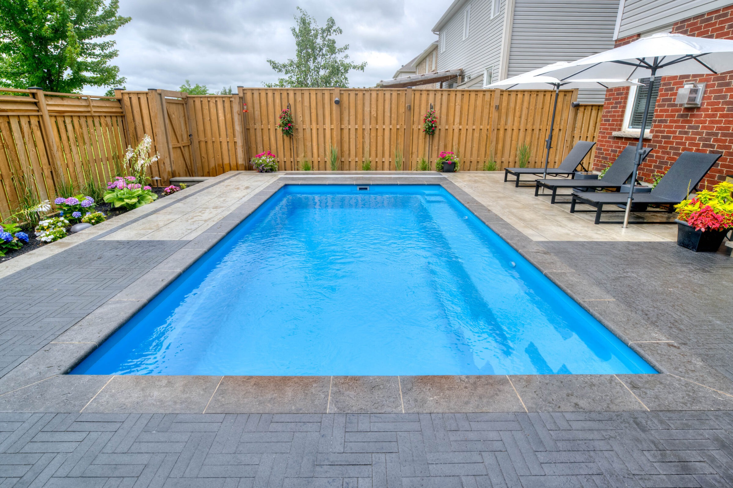 A backyard scene with blue rectangular pool, loungers, umbrellas, and vibrant flowers, enclosed by a wooden fence and house siding.