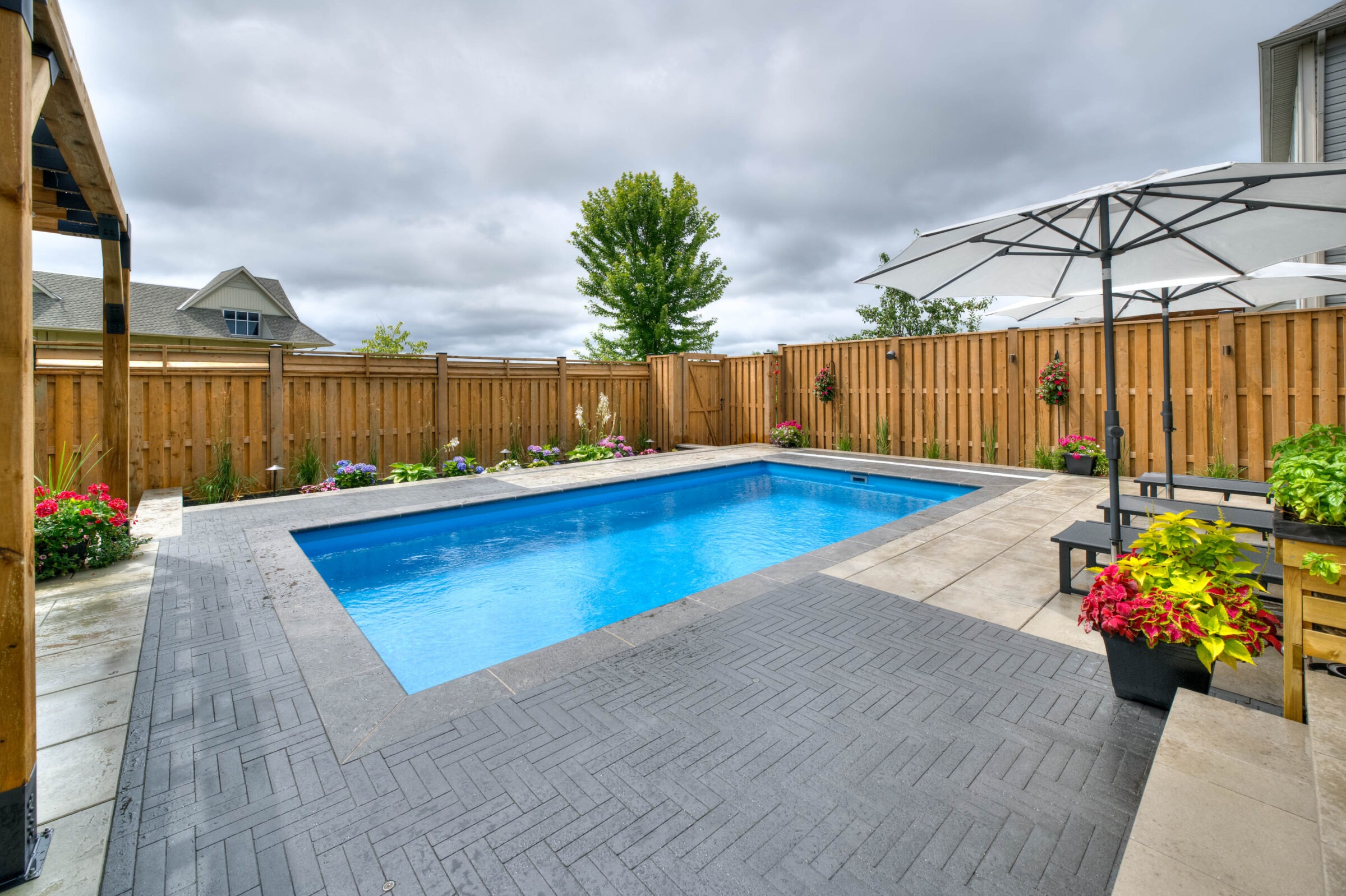 A fenced backyard features a rectangular swimming pool, surrounded by umbrellas, patio furniture, and colorful potted plants under overcast skies.