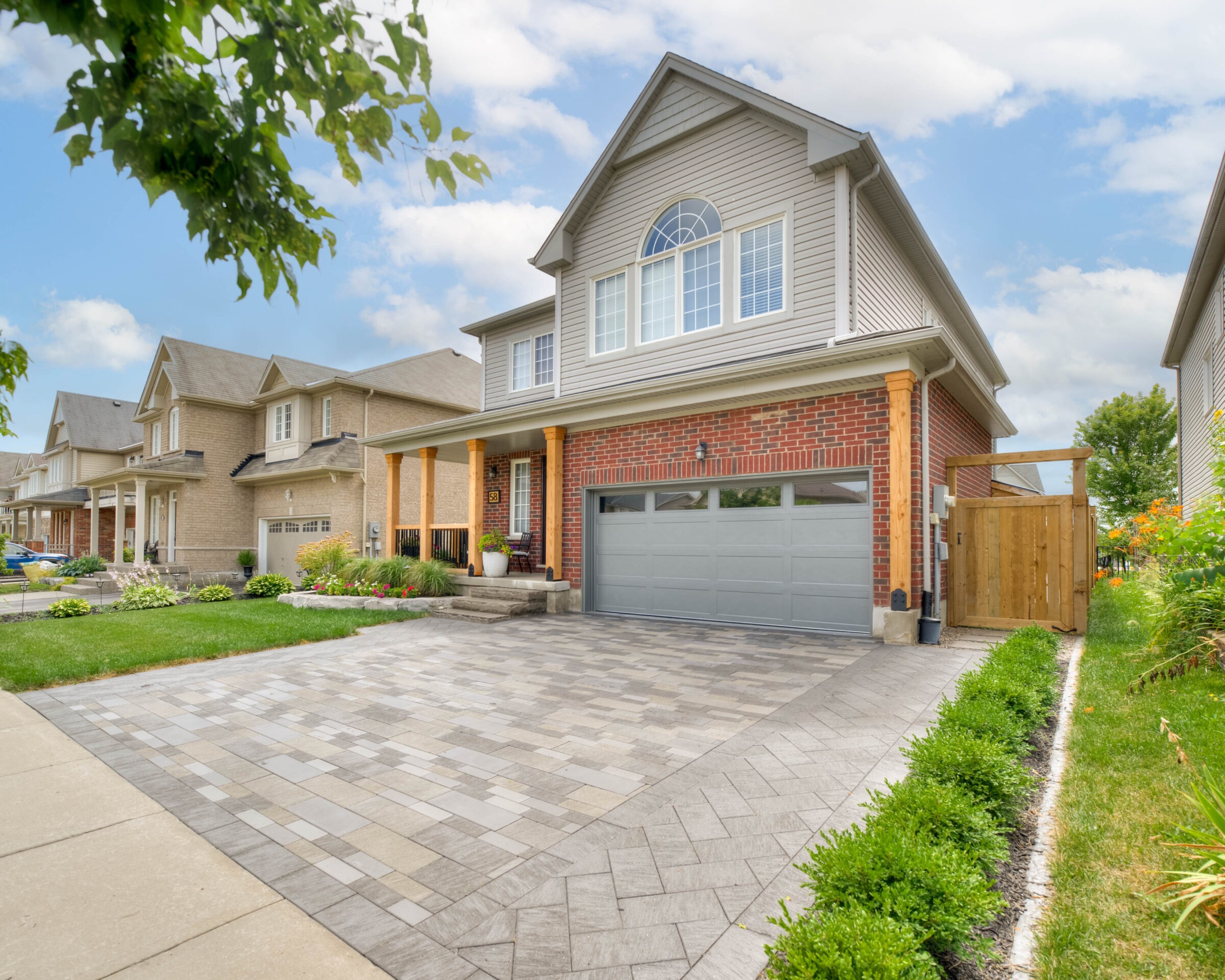 Suburban street with detached houses, featuring red-brick and beige exteriors, landscaped lawns, and paved driveways under a blue sky with scattered clouds.