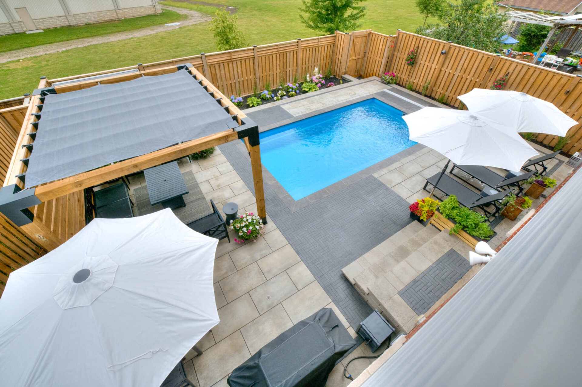 A backyard features a pool, pergola, lounge chairs, and large umbrellas, surrounded by a wooden fence and vibrant garden.