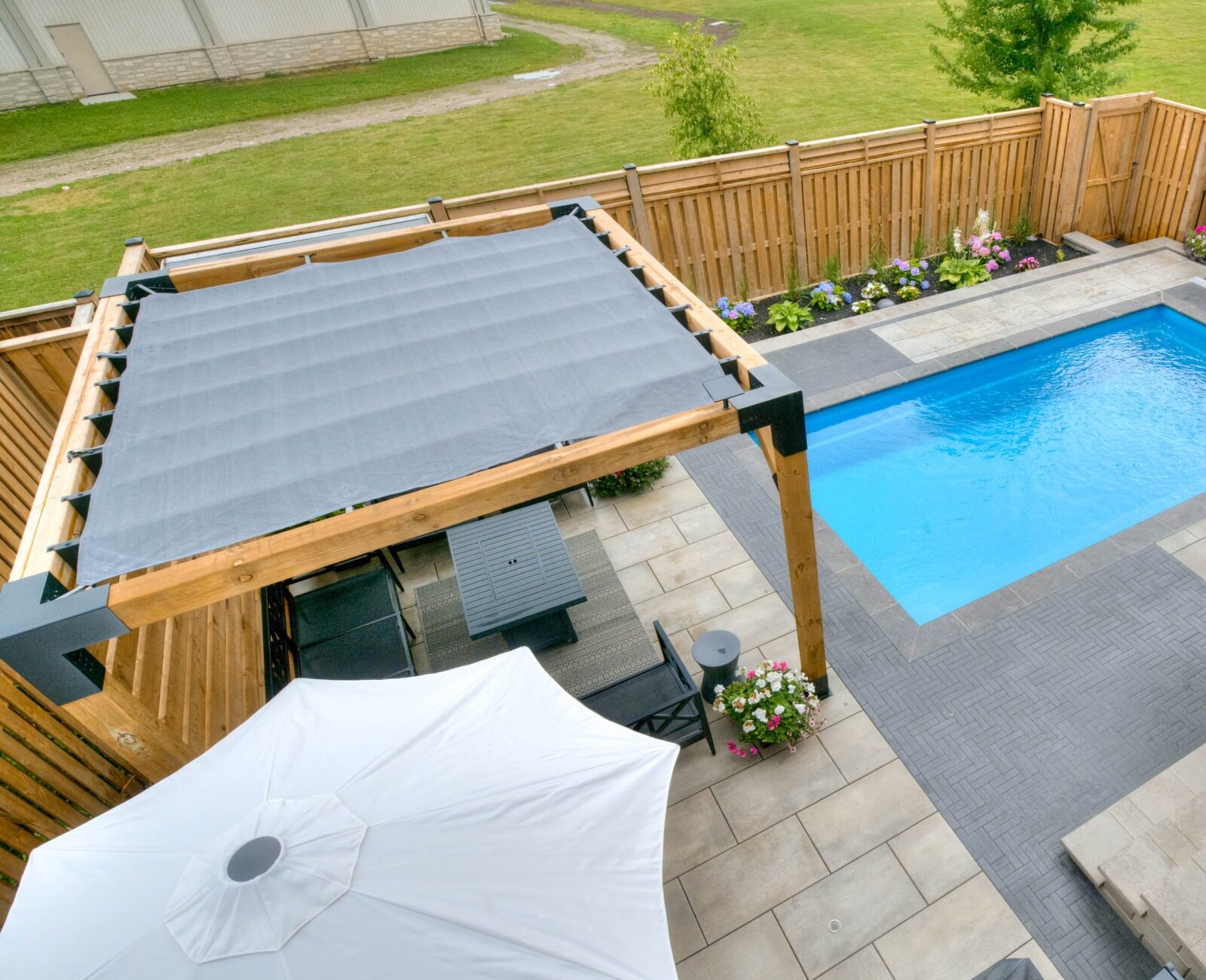 A backyard features a pool, pergola, lounge chairs, and large umbrellas, surrounded by a wooden fence and vibrant garden.