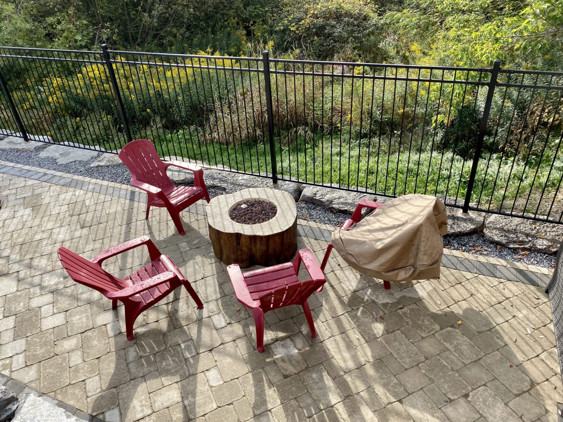 A patio with red chairs arranged around a covered fire pit, enclosed by a black metal fence, overlooking greenery.
