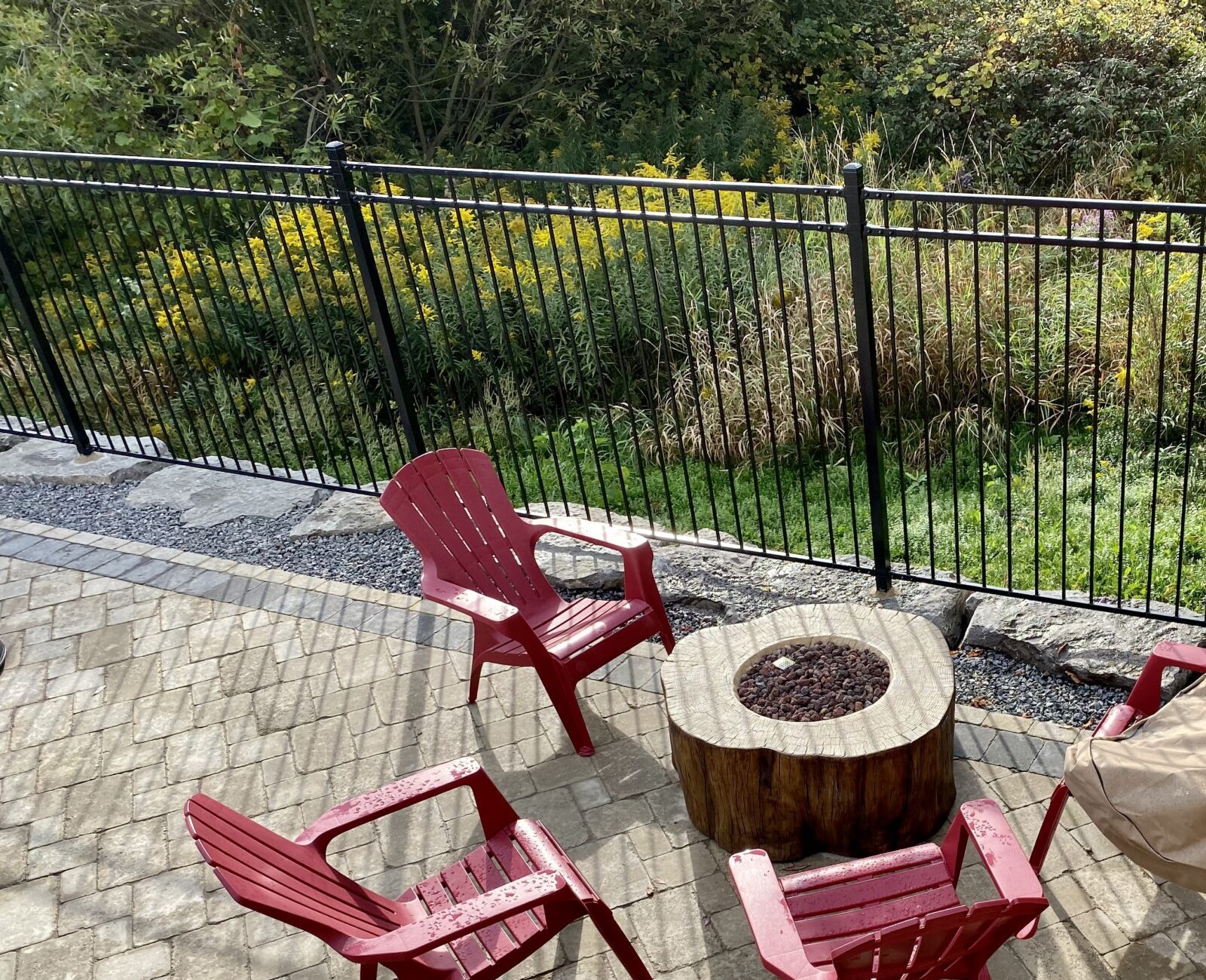 A patio with red chairs arranged around a covered fire pit, enclosed by a black metal fence, overlooking greenery.