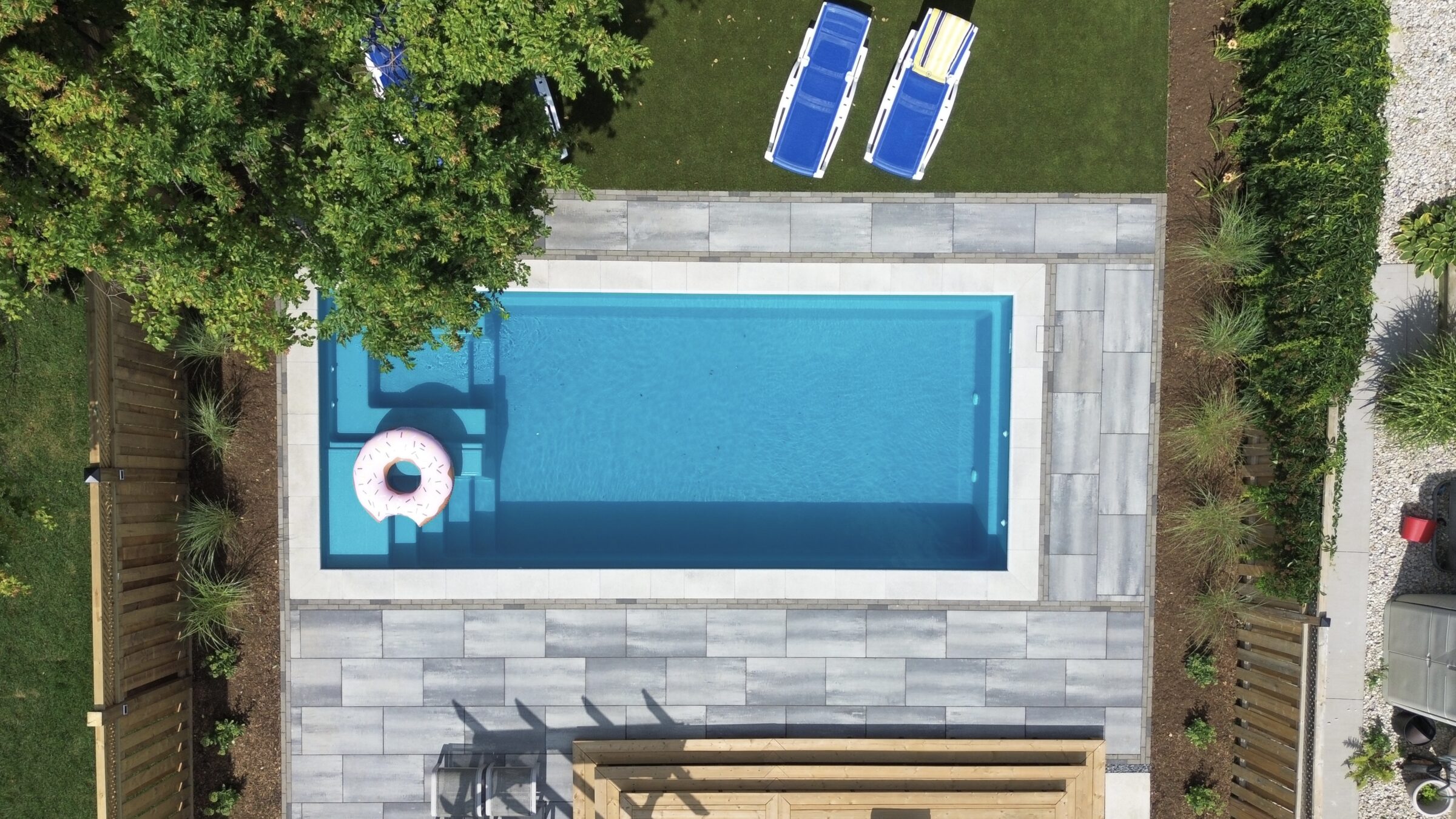 Aerial view of a backyard pool with a donut float, surrounded by lawn chairs, trees, and wooden fencing.