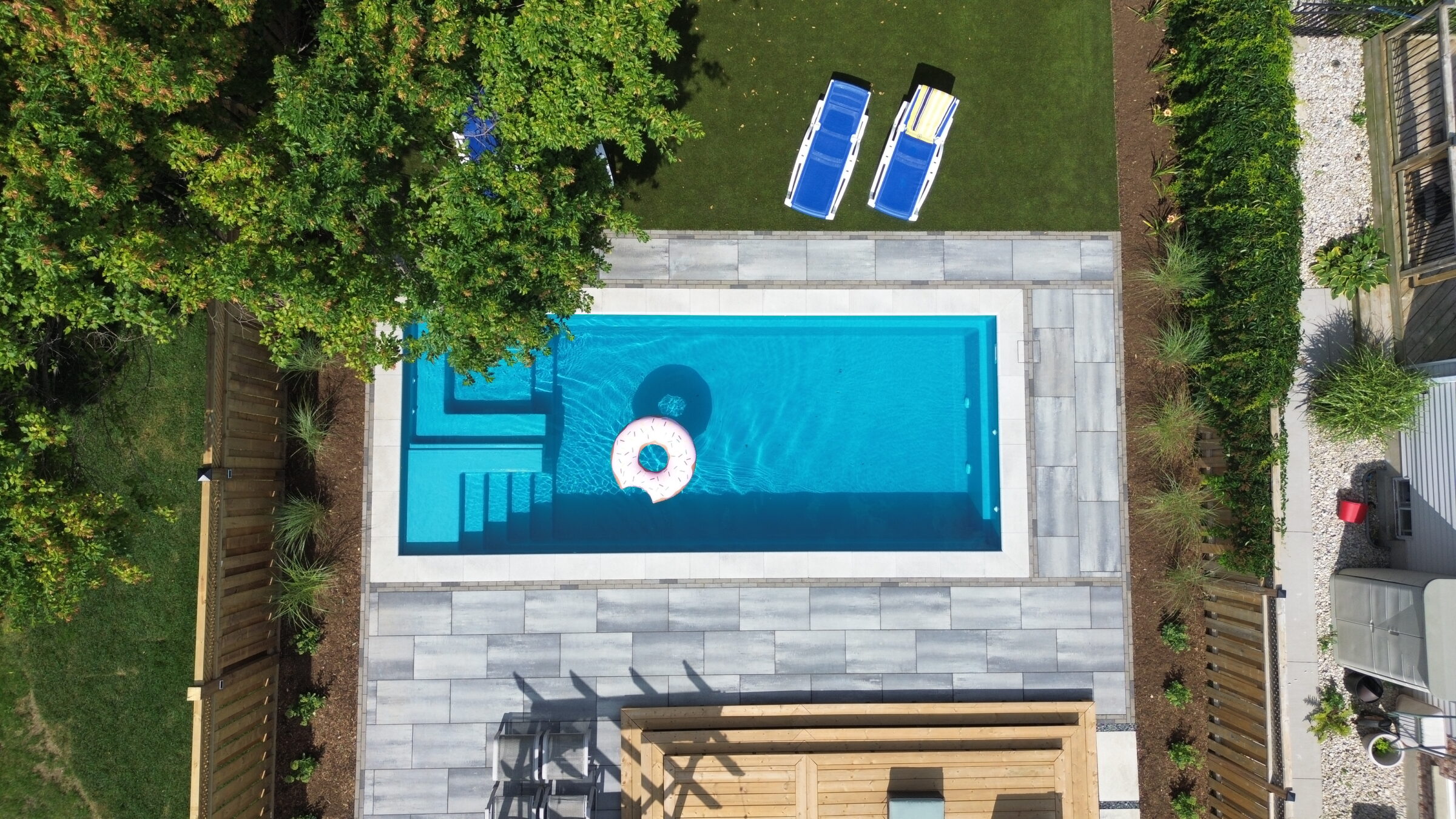 Aerial view of a backyard with a swimming pool, inflatable ring, and two blue lounge chairs on green grass. Wooden deck and greenery surround.