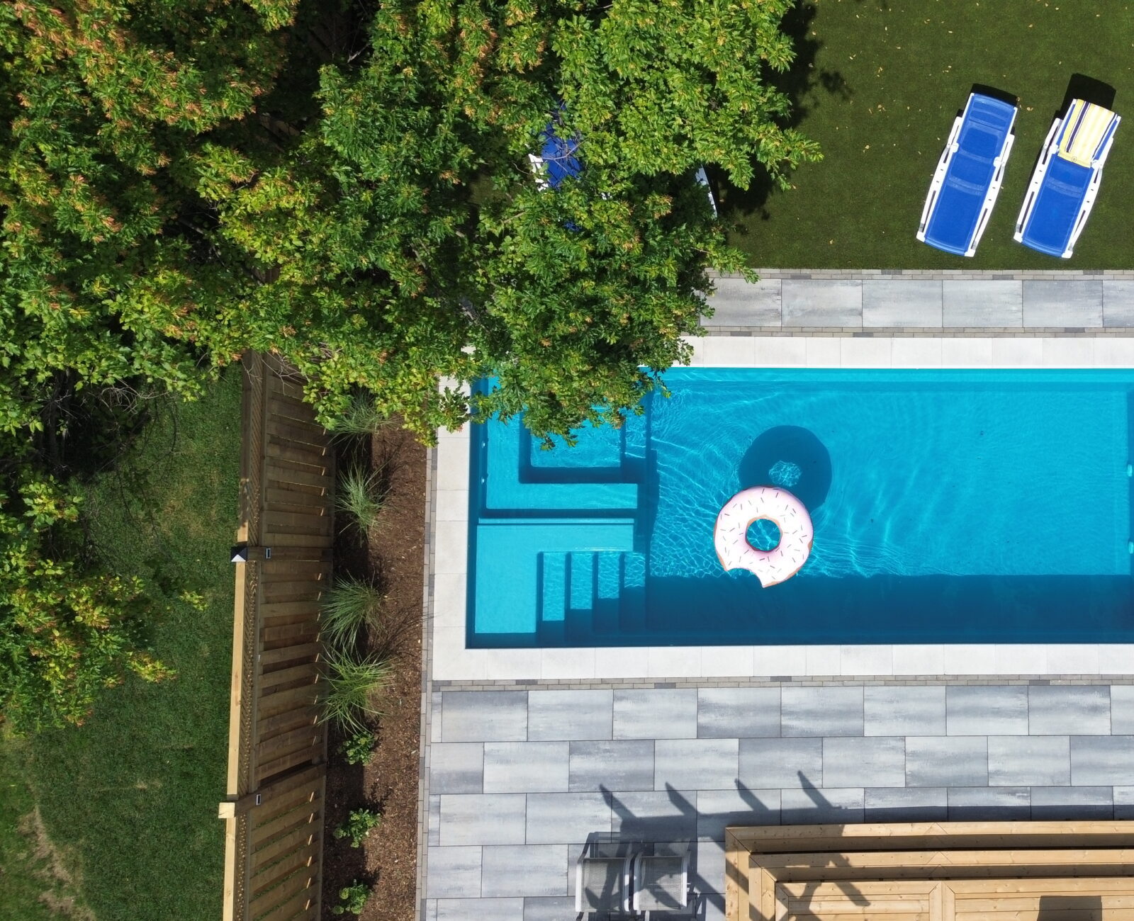 Aerial view of a backyard pool with a float and loungers, surrounded by greenery and a wooden deck area.