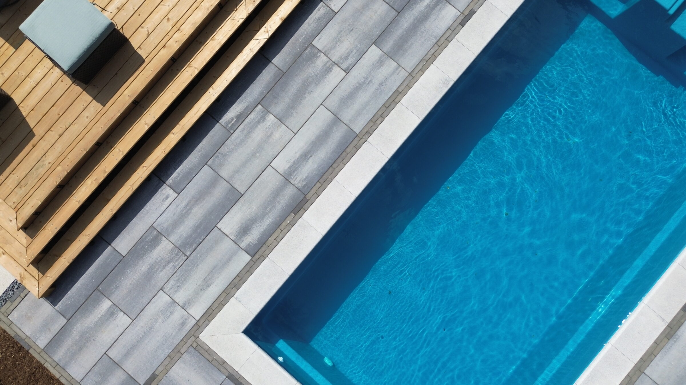 A modern stone patio leads to a clear blue swimming pool, surrounded by wooden decking with cushioned seating, creating a relaxing outdoor area.