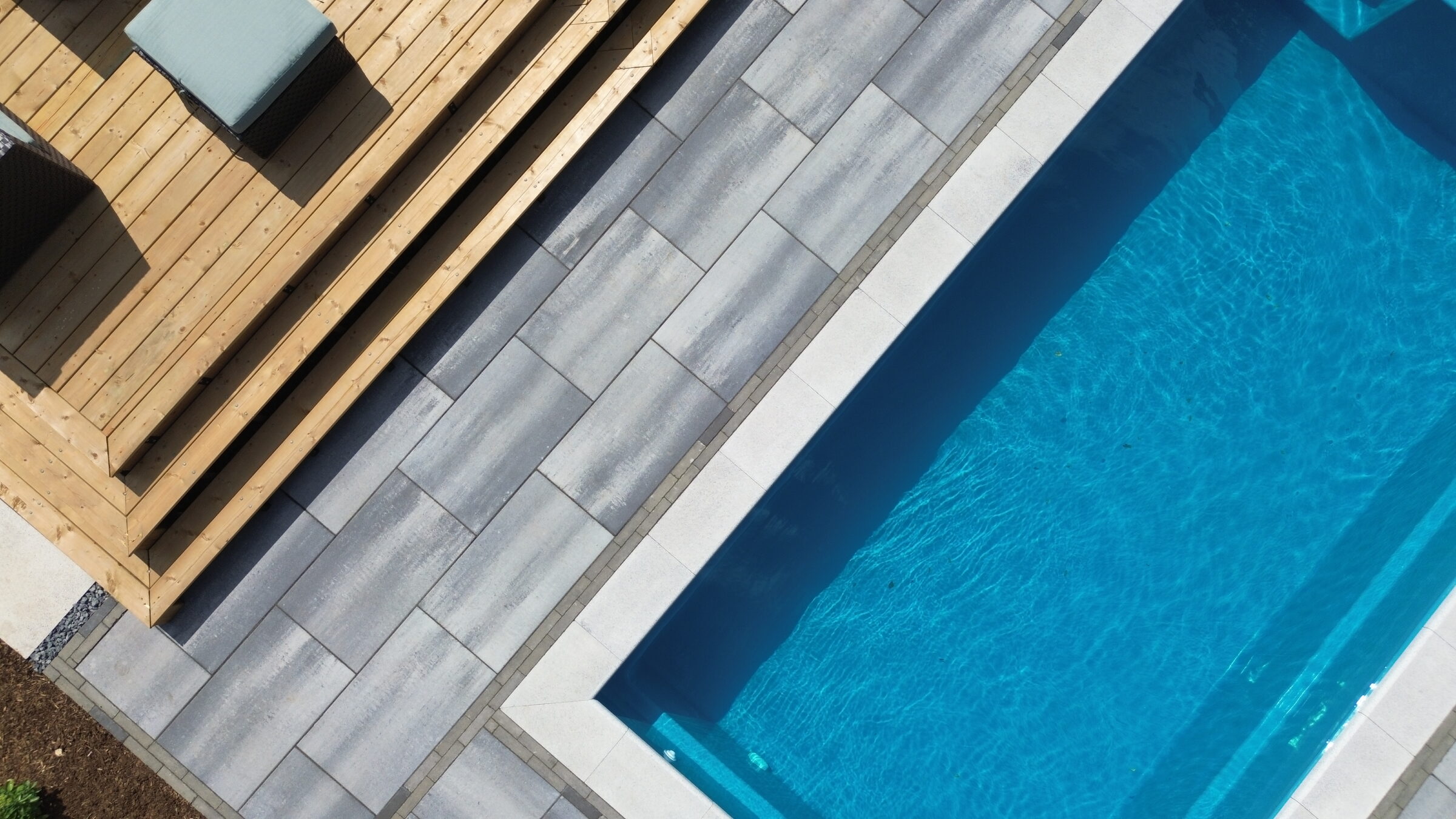 Modern outdoor scene with a blue swimming pool, wooden deck, and gray tiles. Sofas are positioned on the decking area for relaxation.