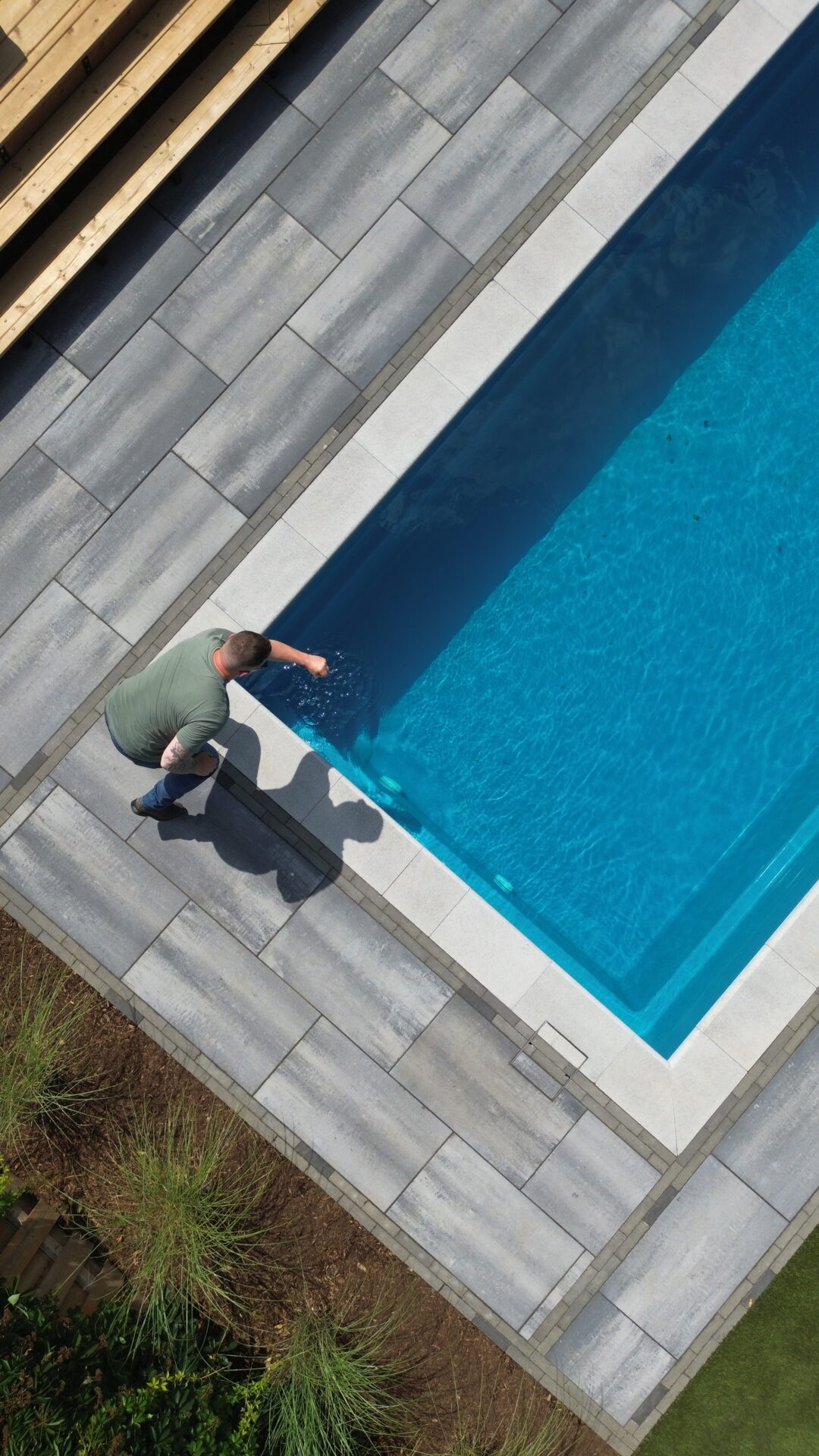 A person leaning over an outdoor swimming pool, surrounded by gray tile flooring and greenery, casting a shadow on the ground.