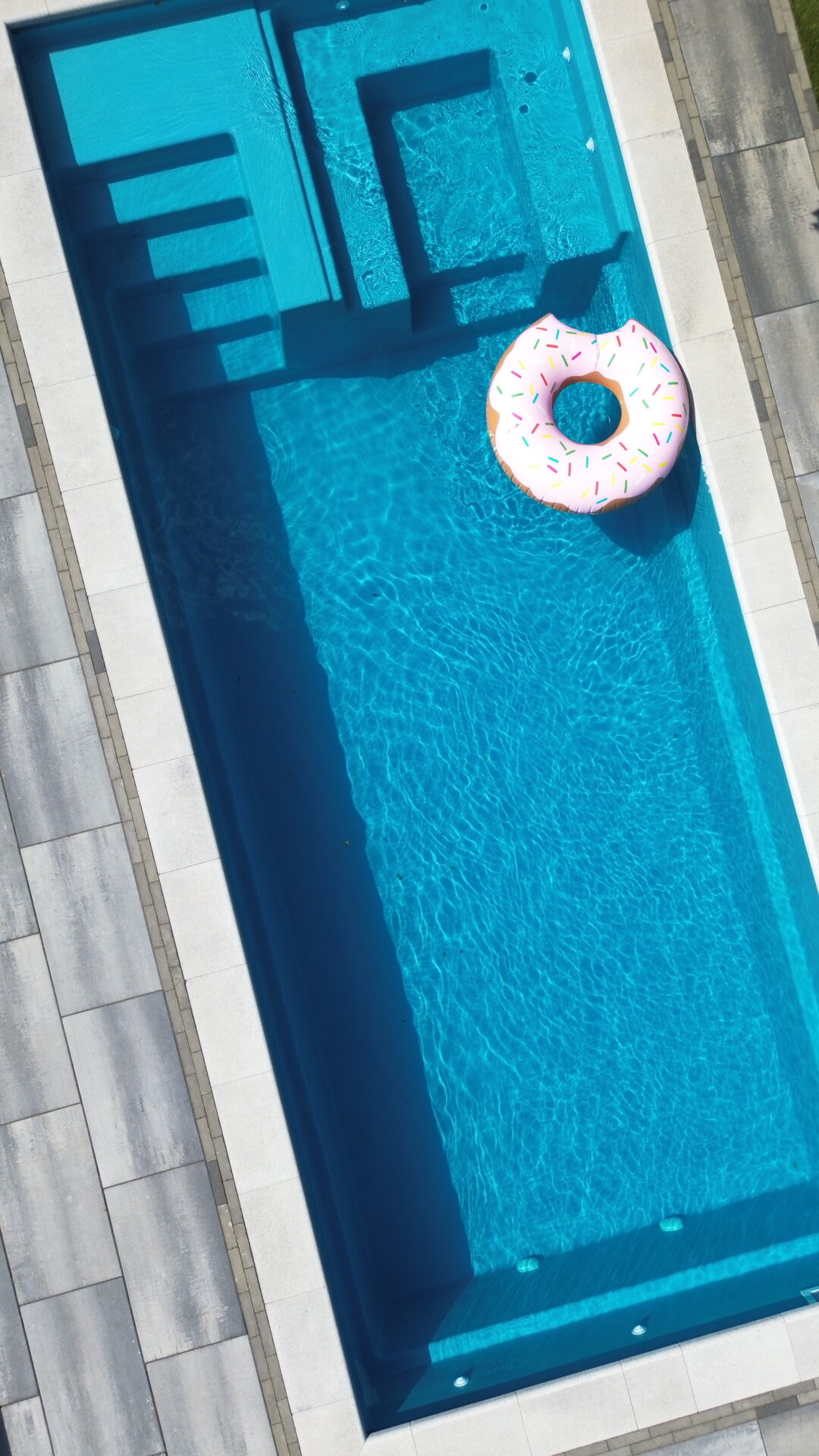 Rectangular swimming pool with clear blue water, surrounded by light tiles. A pink donut-shaped inflatable float rests on the surface.
