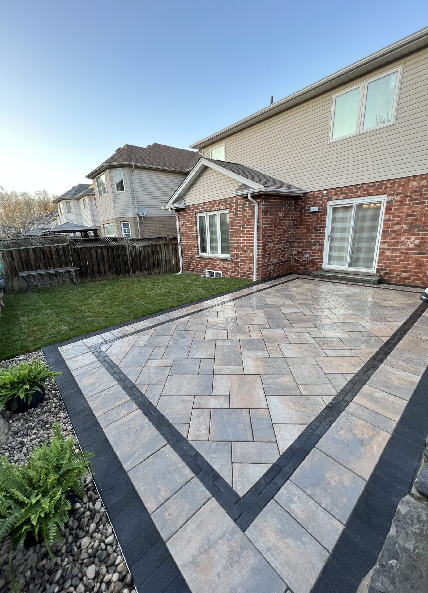 Brick house with modern patio, surrounded by green grass and a wooden fence. Clean, geometric paving stones and a clear blue sky.