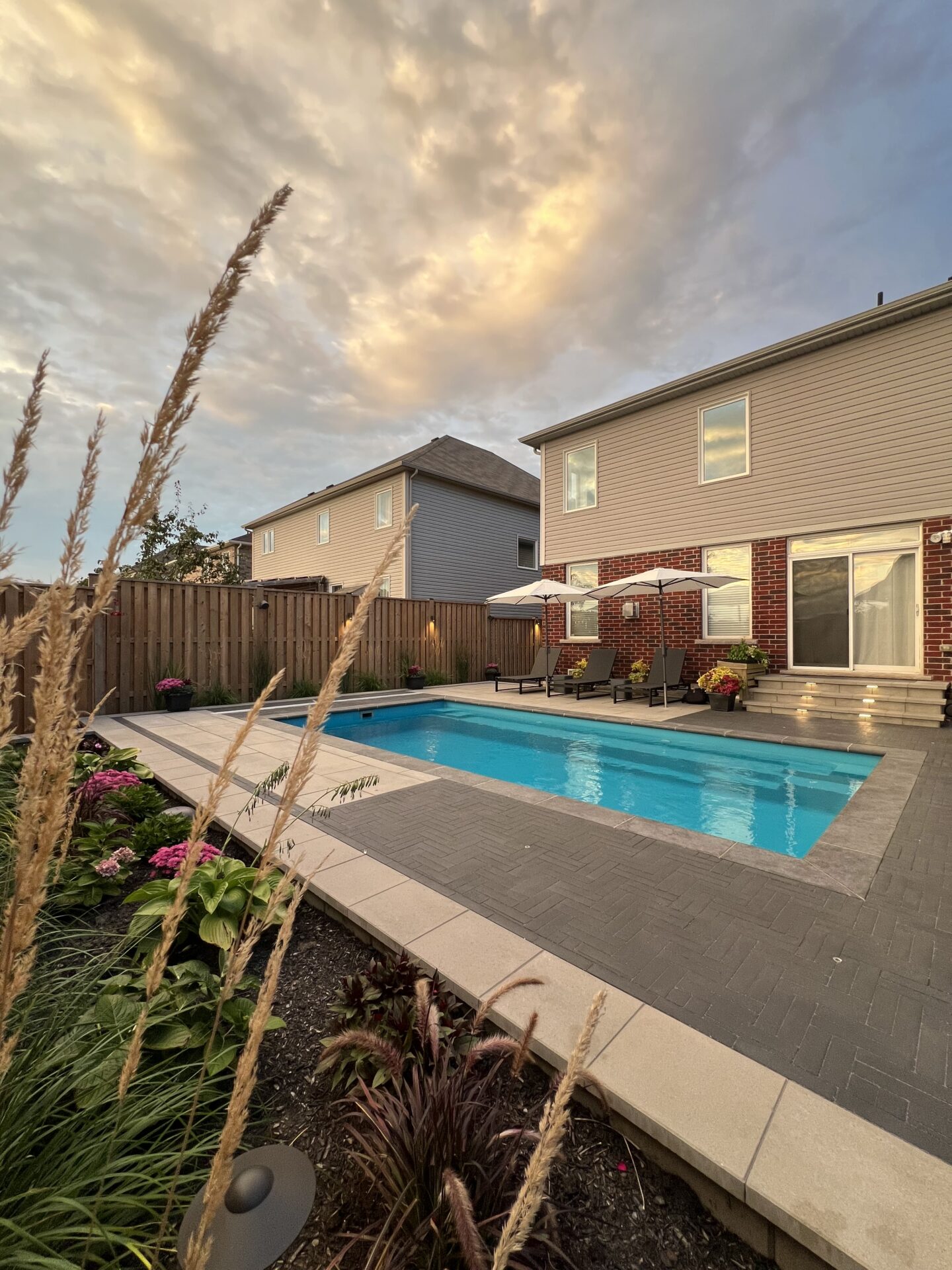 A modern backyard features a rectangular pool, patio furniture, and vibrant plants against a cloudy sky, framed by neighboring houses and wooden fencing.