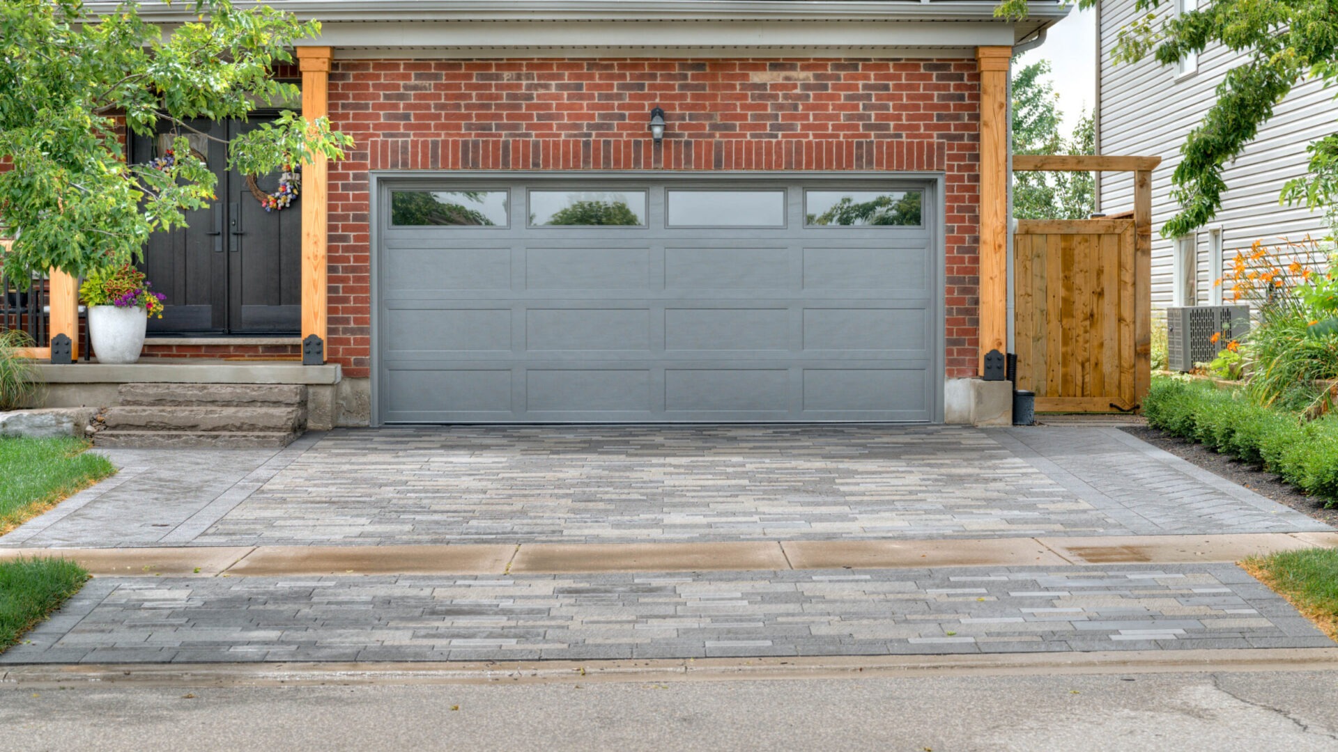 Brick house with a dark gray garage door, modern driveway, and wooden porch. Potted plants decorate the entrance, surrounded by greenery.