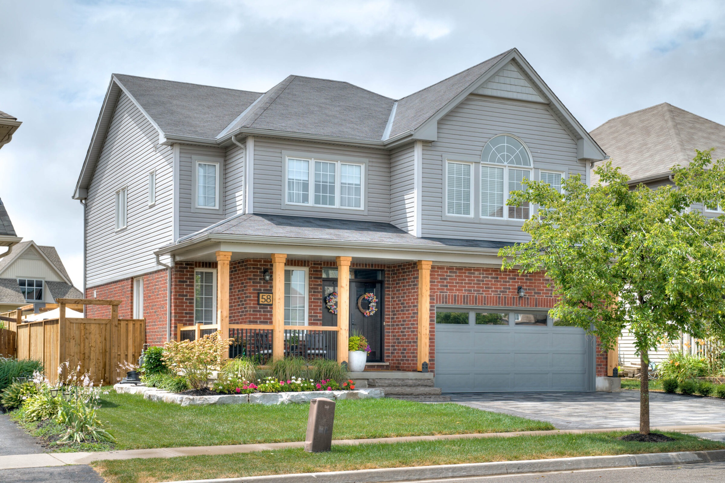 A two-story suburban house with a brick facade, attached garage, porch with wooden pillars, decorative plants, and a neatly maintained front yard.