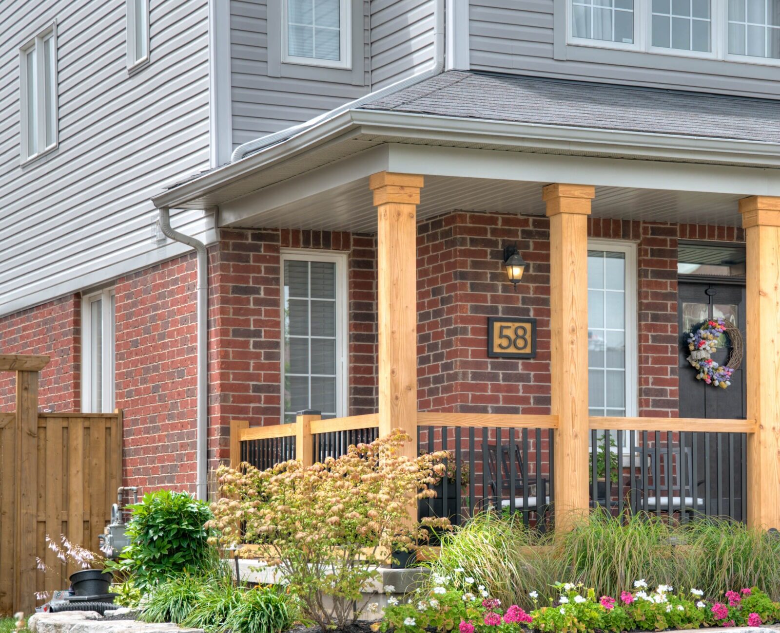 A suburban brick house with a wooden porch, colorful wreaths on the door, surrounded by neatly arranged flowers and green lawn.