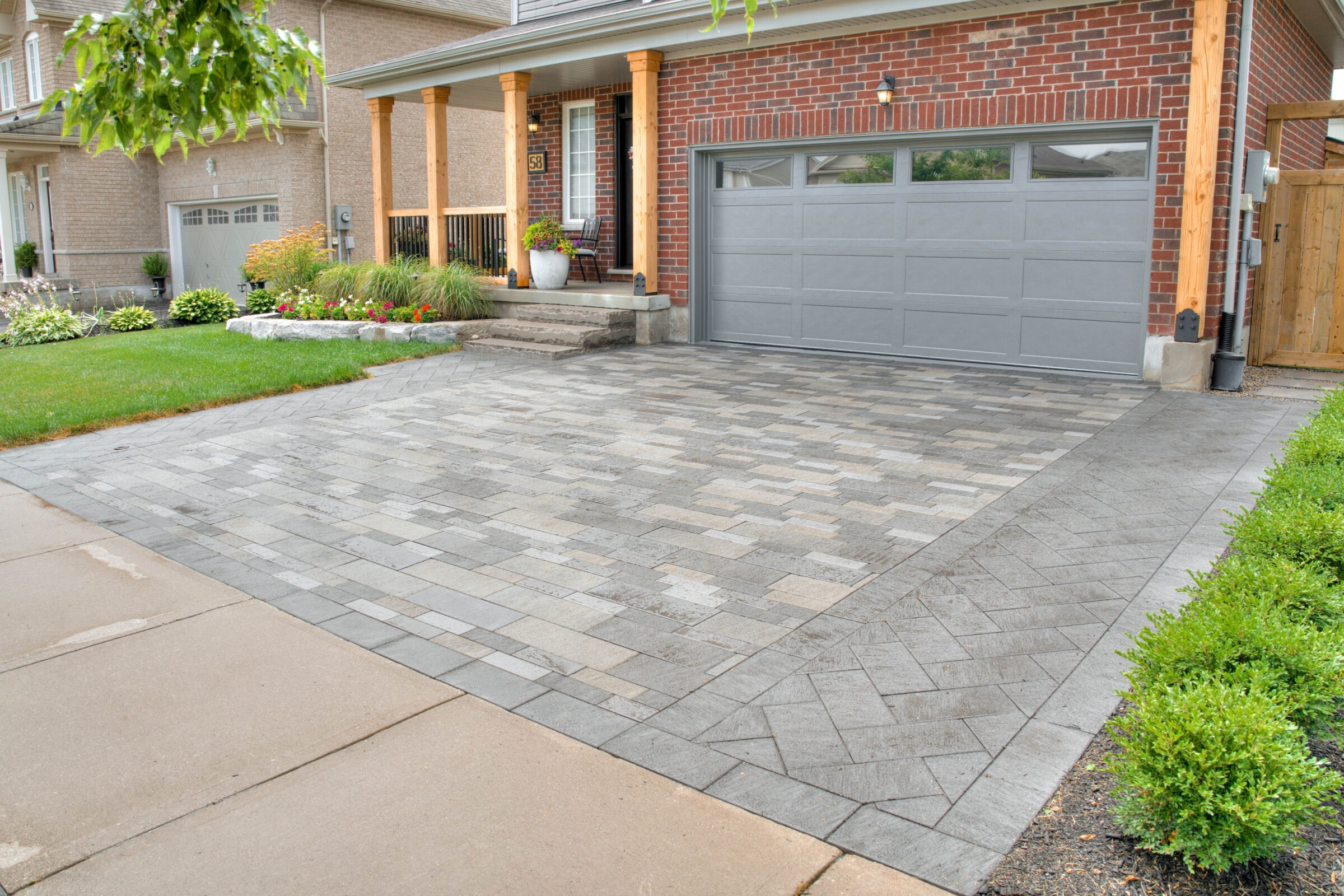 A brick house with a grey garage, landscaped garden, and patterned driveway. Porch with chairs and potted plants complete the inviting scene.