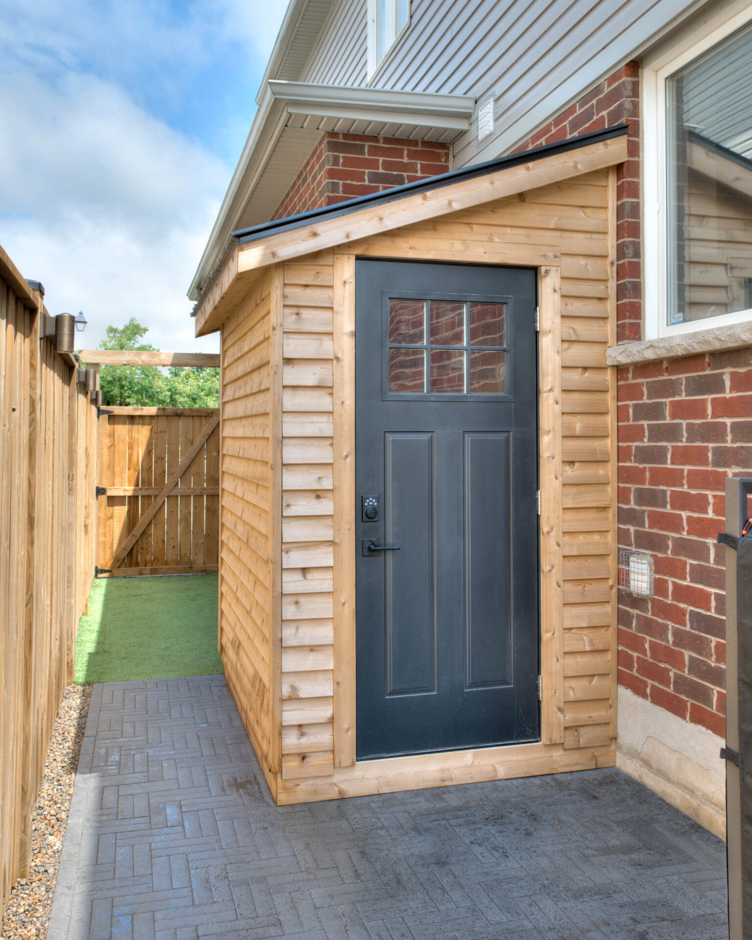 A wooden shed with a black door is attached to a brick house, surrounded by a fenced yard and paved walkway.