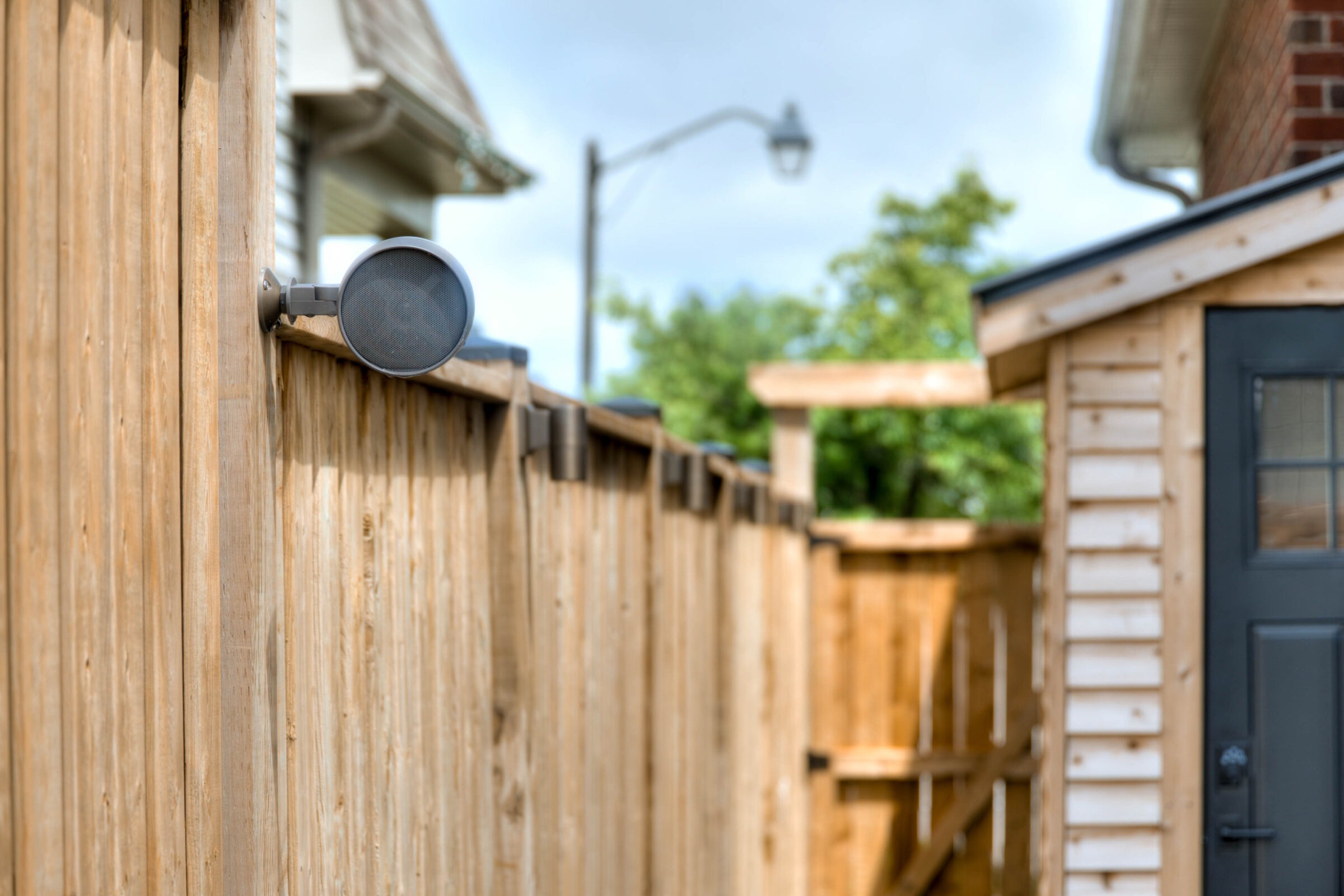 Wooden fence with mounted speaker, next to a small wooden structure. Streetlamp and trees in the background under a clear sky.